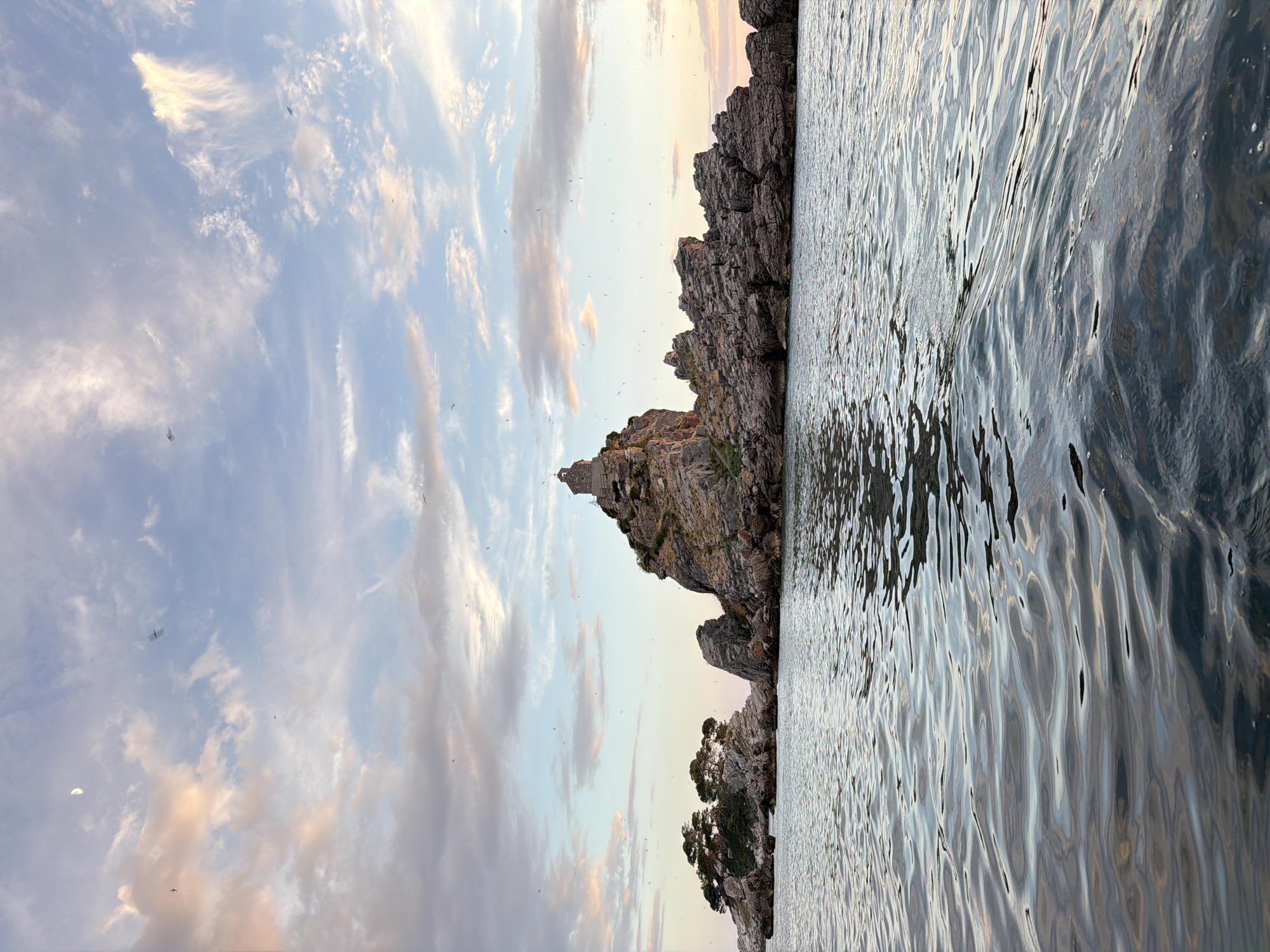 This media file shows a view of the sea and a rocky coastline. The scene is likely Petrovac na Moru, Montenegro, given the location context. The sun is low in the sky, indicating it is either sunrise or sunset, with warm hues tinting the clouds. The water is slightly rippled, reflecting the light from the sky and the rocks. The rocky shore is rugged and appears to be covered in sparse vegetation in places. Small birds are visible in the sky, and a sliver of a crescent moon can be seen in the upper left quadrant of the image. The overall mood is serene and natural. There is no visible text in the image.