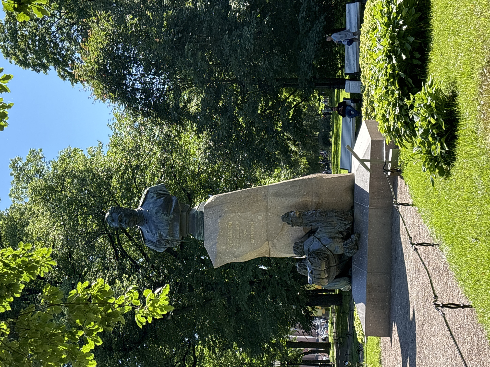 An outdoor, eye-level, medium shot captures a bronze bust of a man in military uniform, mounted on a large, irregular stone pedestal. The pedestal features inscribed text and two bronze sculptures of camels at its base, with one camel carrying saddlebags. The statue is situated in a park-like setting with lush green grass and a gravel path in the foreground. Behind the statue, large trees with dense foliage create a natural backdrop, and the clear blue sky is visible through gaps in the canopy. In the middle ground, several white park benches are occupied by people, including a man seated with a mobile phone and a woman standing. Sunlight filters through the trees, casting dappled shadows on the ground. The overall scene depicts a monument in a public park on a sunny day.