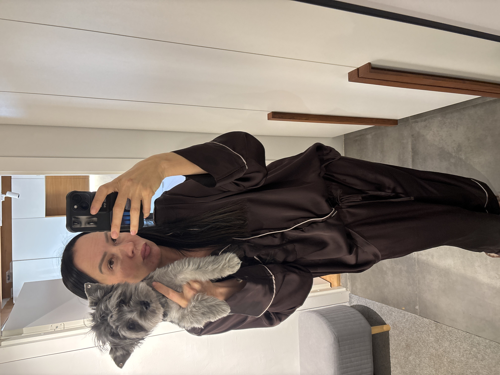 The image appears to be a selfie taken in an indoor setting, likely a residential space.
**People:**
* A woman is holding a small dog. She is taking the photo with a phone. She is wearing dark-colored pajamas with white piping.
**Subjects:**
* The small dog is a focal point in the photo. It is a grey color.
**Objects:**
* A smartphone is held up to capture the image.
* A piece of furniture appears in the bottom left of the image.
* The interior includes doors and walls with wooden elements.
**Actions:**
* The woman is taking a selfie.
* The dog is held by the woman.
**Interactions:**
* The woman is interacting with the camera (taking the photo) and the dog (holding it).
**Environment:**
* The setting is an interior space, possibly a room in a house or apartment. The walls are white.
* Based on visual evidence in the background, there is no definitive indication of the city of Chaikovsky, Russia.