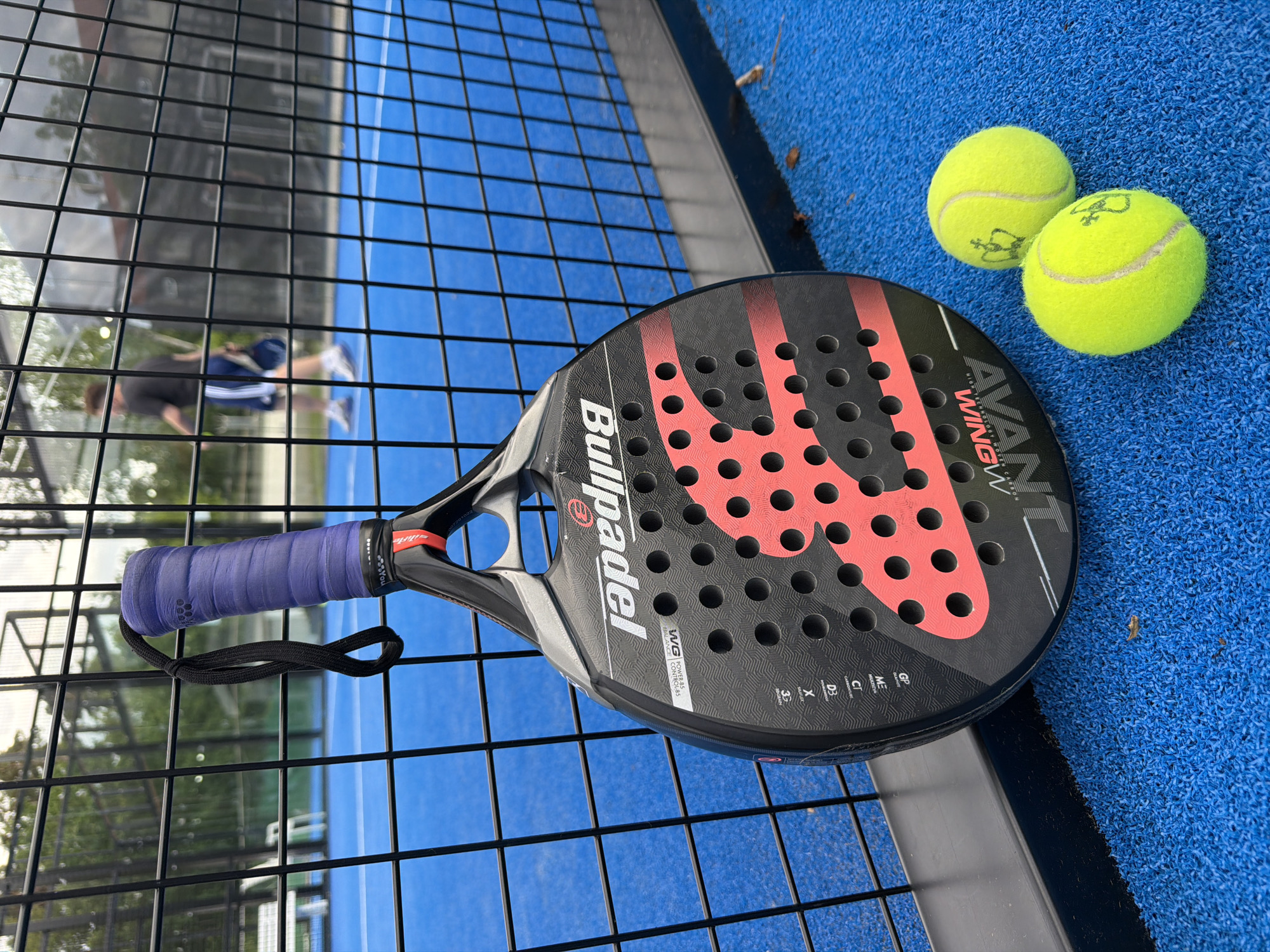 A padel racket and two balls are positioned on a blue padel court, with a black metal fence in the foreground and background. A person, out of focus, walks in the background of the court. The racket is black with red accents and has the brand name "Bullpadel" visible. The balls are yellow and fuzzy, with a crown logo. The scene suggests a sporty, outdoor setting, likely a recreational facility. The image was taken in Vaterstetten, Germany.