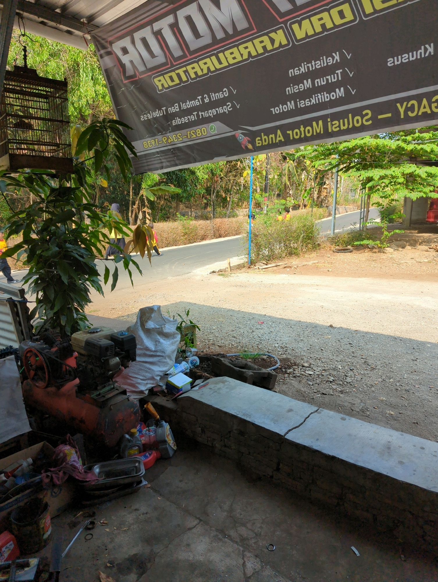 The image depicts an outdoor workshop or repair shop, likely located in Kedungdowo, Indonesia. A large banner with text in Indonesian and English is prominently displayed. The text on the banner mentions "MOTOR DAN SPAREPART" and "SOLUSI MOTOR Anda". Below this, a list of services appears to be provided, including "Kelistrikan", "Turun Mesin", "Modifikasi", and "Perbaikan". A phone number, "0895-6405-1580", is also visible.
In the foreground, there is a red air compressor and other mechanical equipment, suggesting it is an engine repair shop. Various tools, oil cans, and a metal tray are scattered around, indicating an active work area. A birdcage hangs from the ceiling to the left of the banner.
In the background, a paved road is visible, with trees and vegetation lining its sides. Several people are walking on the road, some wearing bright clothing. The overall environment appears to be a rural or semi-urban setting. The lighting suggests it is daytime, with sunlight casting shadows. The ground surface consists of gravel and concrete.