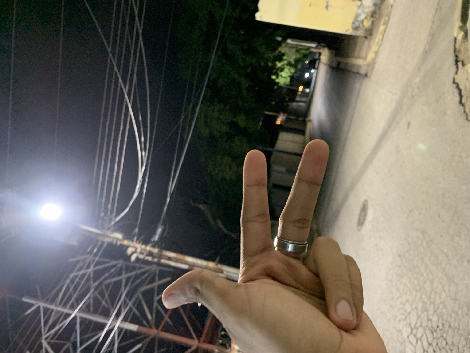 The image depicts a nighttime street scene in Santa Ana, Venezuela. In the immediate foreground on the left, a human right hand, exhibiting a medium-dark skin tone, is prominently positioned. The thumb is extended upwards, while the index and middle fingers are extended horizontally to the right, forming a "V" shape, commonly known as a peace sign or indicating the number two. A silver-colored, band-style ring is worn on the ring finger.
The ground beneath the hand consists of a light-colored, patterned pavement, likely a sidewalk, which transitions into an asphalt street. A circular manhole cover is visible on the street surface in the lower mid-frame.
In the midground, a brilliant, circular street lamp illuminates the scene from the upper center, positioned above a utility pole. Numerous utility wires are stretched across the dark sky, connecting to the utility pole and other unseen structures. To the left and slightly behind the hand, a large, intricate metal lattice structure, possibly part of a communication tower or significant utility support, is discernible. The asphalt street recedes into the background, flanked by a yellow curb on the left side and a building facade with faded yellow paint and visible weathering on the right.
The background features dark green foliage of trees lining the street. Faint, blurred lights, consistent with vehicle headlights or taillights, are visible further down the road, indicating the presence of distant vehicles. The overall sky is dark, consistent with a nighttime setting.