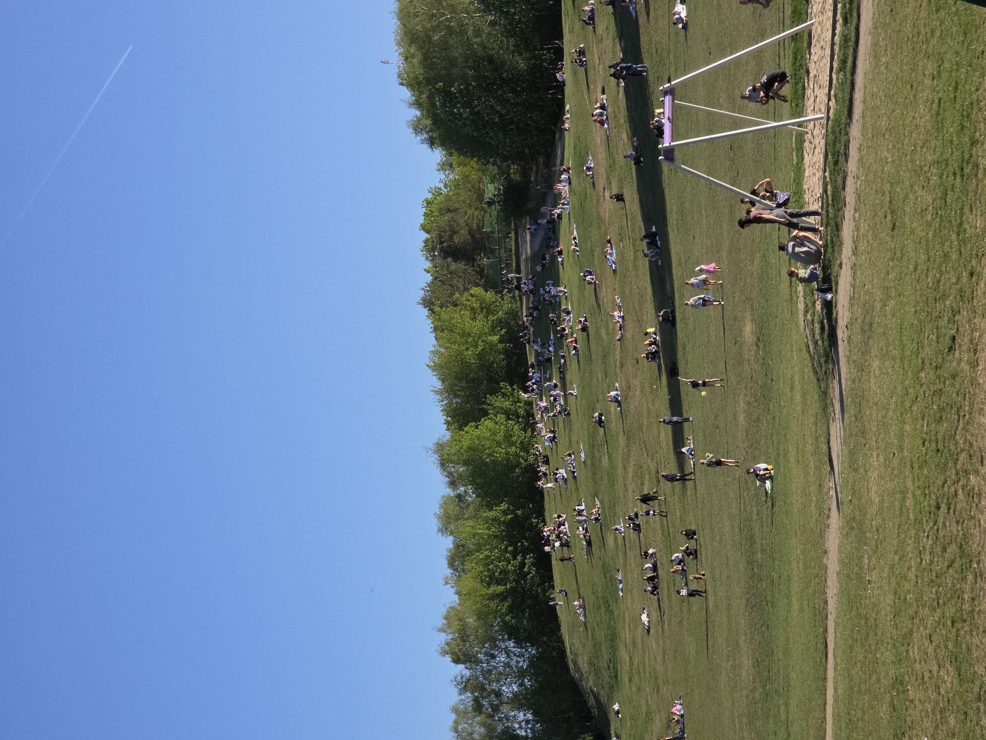 A large grassy field is full of people enjoying a sunny day. The field is bordered on one side by trees and on another side by a sandy area and what appears to be a structure or playground equipment. People are spread out across the field, some lying down, some standing, some in groups. The sky is clear and blue, with a contrail visible in the upper left. The location is most likely Pole Mokotowskie in Warsaw, Poland.