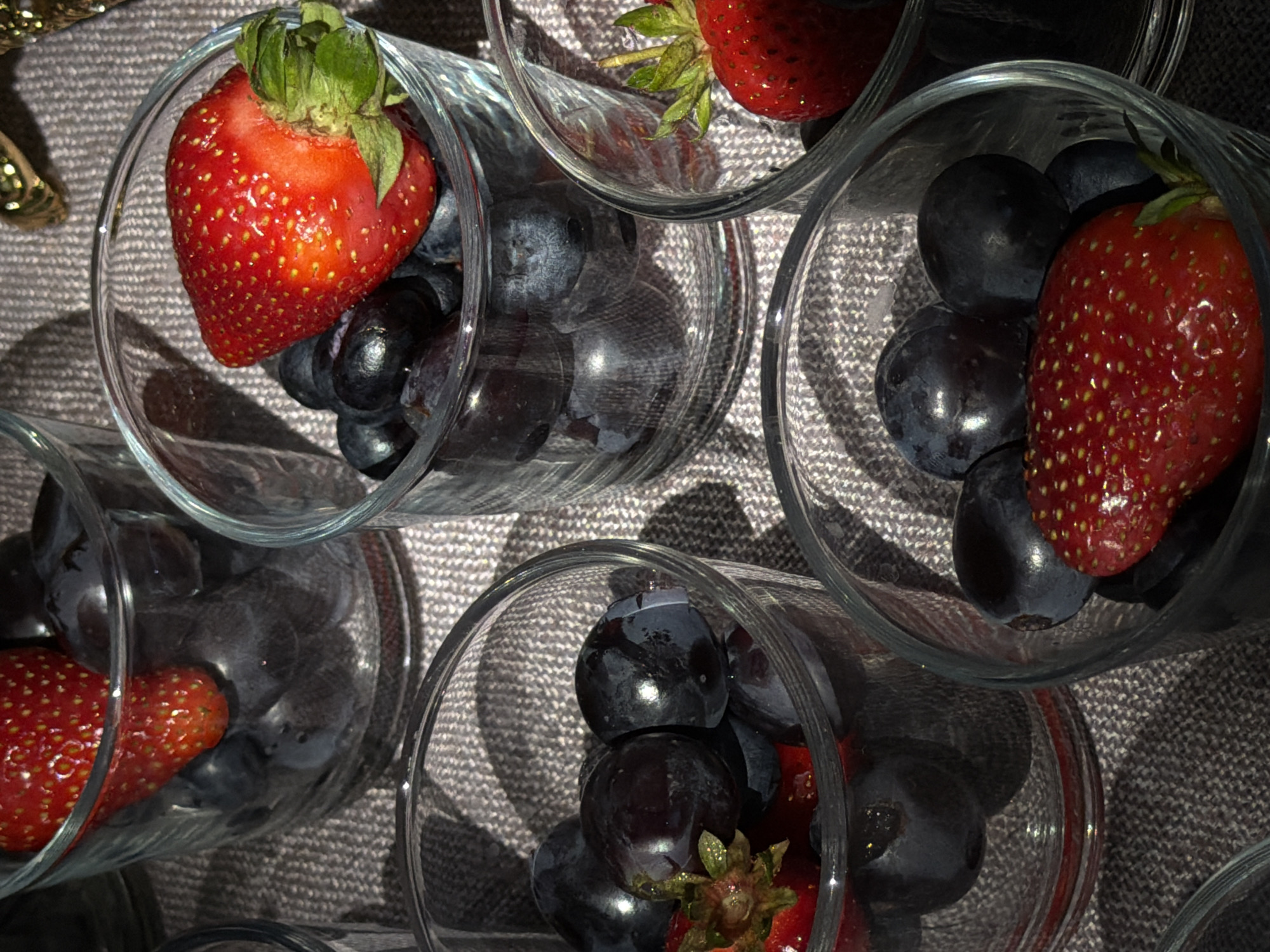 The image shows an overhead shot of several clear glasses filled with blueberries and strawberries. The glasses are arranged close together on a textured grey surface that appears to be a cloth. Each glass contains a mix of dark blueberries and one or two bright red strawberries with green tops. The strawberries and blueberries vary in size and arrangement within each glass. The lighting suggests an indoor setting with a light source from above, casting shadows and highlights on the fruit and glass surfaces. No people are visible. There is no evidence that this image was taken in Bishkek City, Kyrgyzstan.