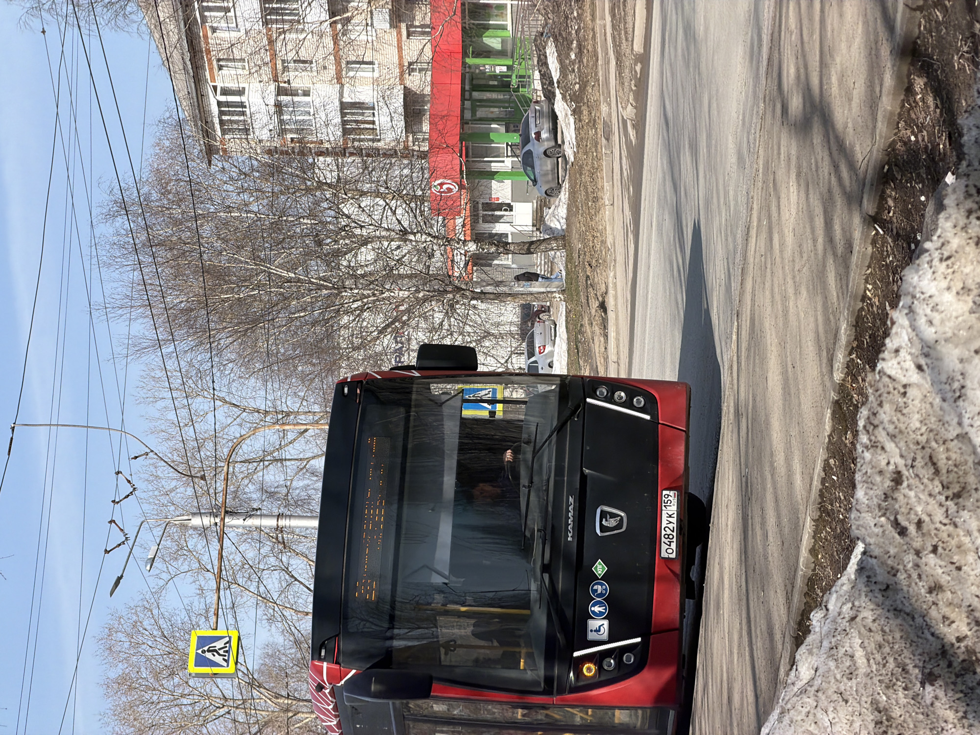 The image shows a red KAMAZ bus with license plate O482YK159 parked on a street in Berezniki, Russia. A pedestrian crossing sign is visible near the front of the bus. A multi-story brick building is behind the bus, and a store or business with red and green signage occupies the ground floor of the building. There is residual snow on the side of the road. Several bare trees are present.