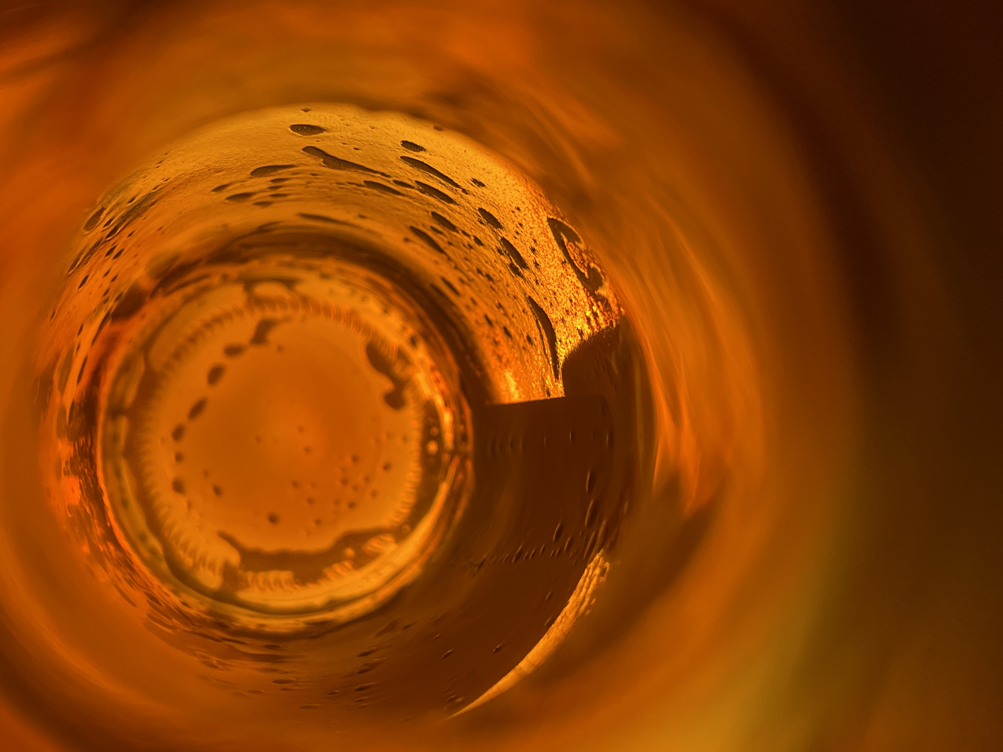 The image shows the interior of a cylindrical container, likely a glass or bottle. The inside surface is coated with a viscous, amber-colored liquid, leaving droplet-like residues and a distinct ring at the base. Light entering the container creates gradients of illumination, with the left side and base appearing brighter. The overall warm tone and swirling patterns within the liquid suggest a perspective looking into the bottom of a partially emptied container. There's no information about the location in the city of Подшивалово, Russia.