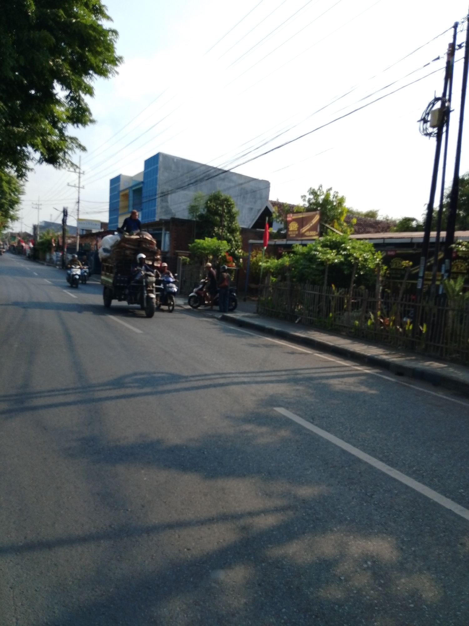 A street scene in Pasuruan, Indonesia, features a multi-lane asphalt road under a bright sky with scattered clouds. Power lines run overhead, supported by utility poles. On the right side of the road, a weathered bamboo fence lines the edge of a property with lush green trees and bushes. A building with a modern design, featuring blue and grey elements, is visible in the background. A truck is laden with what appears to be hay or agricultural produce, with a driver and at least two passengers on top, navigating the road. Several motorcycles are also present, with riders and passengers. The sunlight casts distinct shadows on the road, indicating a sunny day.