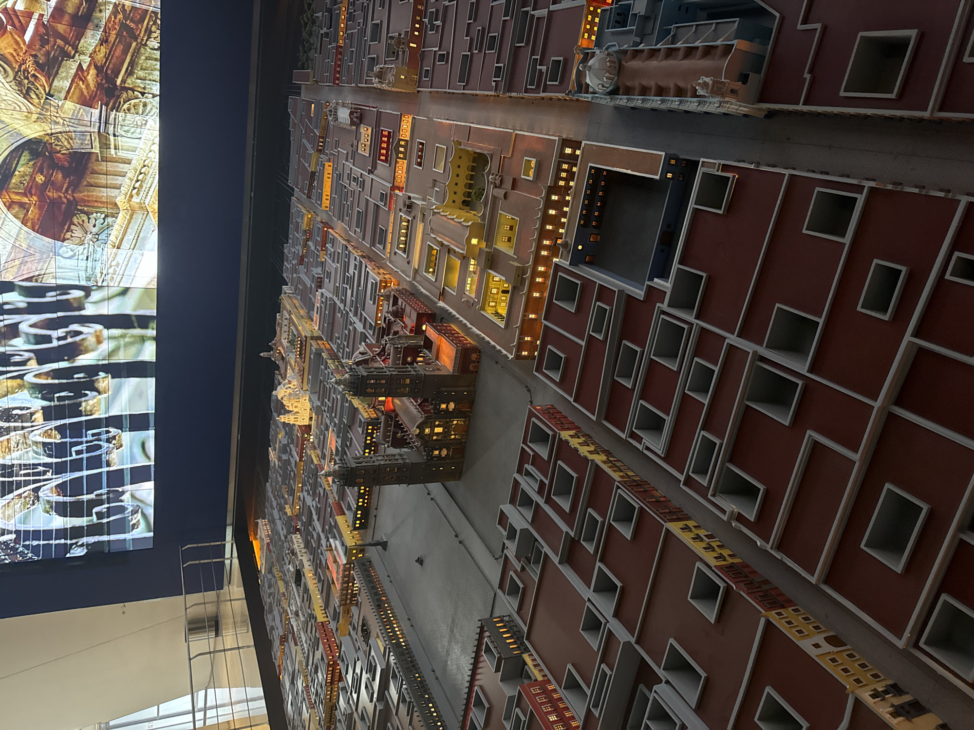 The image shows a detailed architectural model of a city, likely displayed in a museum or exhibition setting. The model consists of numerous buildings, seemingly constructed from wood or a similar material. The buildings are painted in various colors, primarily red, yellow, and gray, and feature repetitive window patterns. Some buildings have interior lights, creating a warm glow. Streets are visible between the buildings, suggesting a grid-like urban layout. To the left of the model is a large screen displaying ornate architectural details, possibly related to the city being modeled. The lighting is dim, highlighting the model's illuminated elements. The city depicted is possibly located in San Bernardino Tlaxcalancingo, Mexico, based on the architectural style visible in the model.