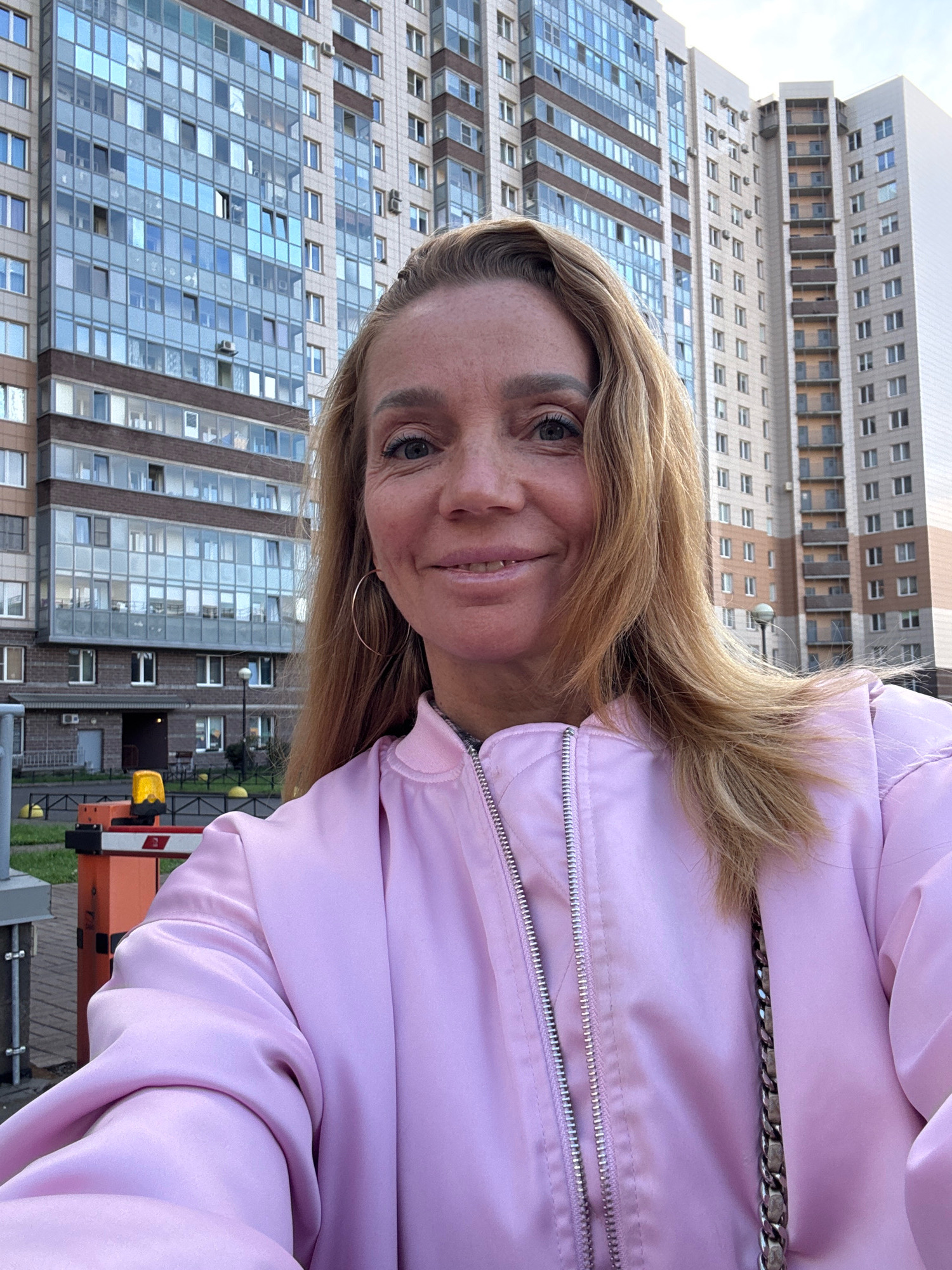 A woman with blonde hair and light-colored eyes takes a selfie outdoors in front of a tall apartment building in an urban environment. She is wearing a pink bomber jacket. A barrier gate with a yellow light is visible in the background, suggesting a residential complex or parking area. The sky is overcast.