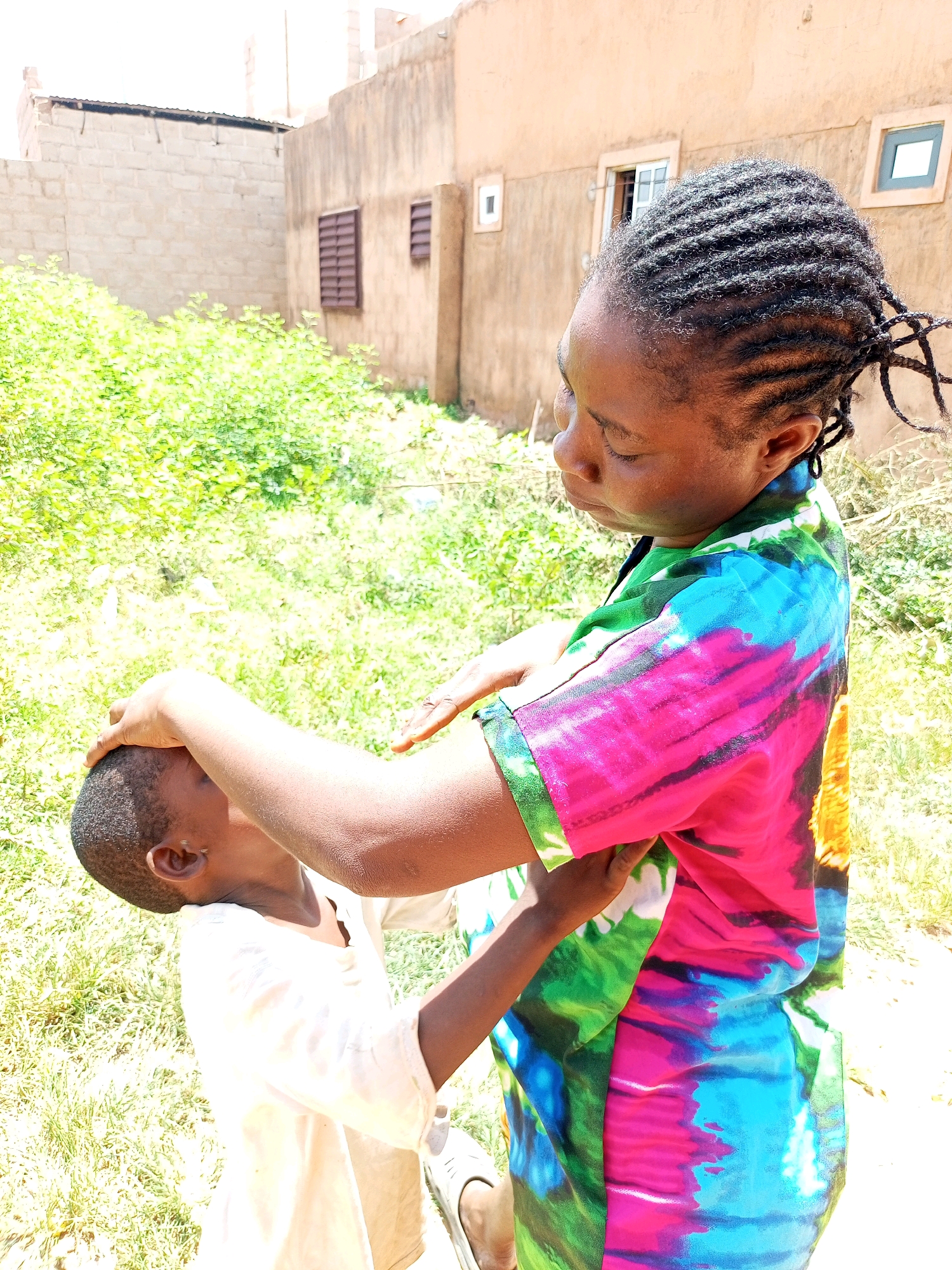 In Tempure, Nigeria, a woman with braided hair is gently tending to a young child outdoors. The woman is wearing a vibrant, multi-colored tie-dye shirt with shades of pink, blue, green, and yellow. She has her hands on the child's head, possibly adjusting their hair or offering comfort. The child is wearing a light-colored shirt and is positioned looking upwards towards the woman. The background shows a slightly blurred environment with green foliage and buildings constructed of brick and a lighter-colored material. Sunlight illuminates the scene, casting shadows and highlighting the textures of the surroundings. The ground is covered in dry grass and dirt.