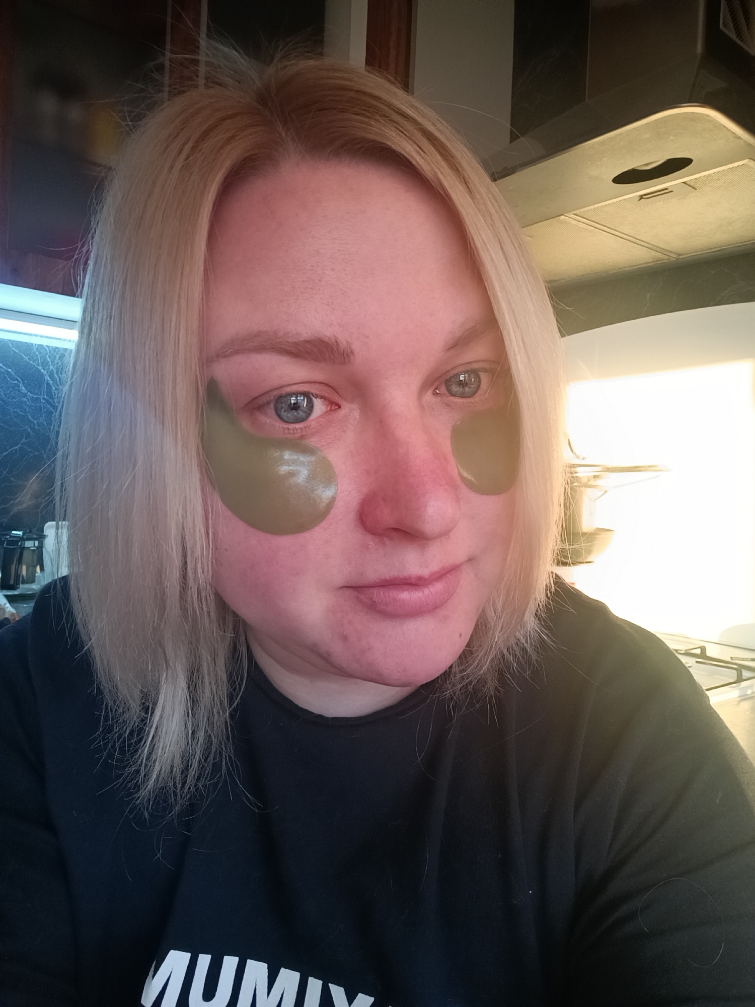 A blonde-haired woman with eye patches is posing for a selfie. She is wearing a black top with white text, partly obscured. The background features a kitchen environment, possibly in a residence in the city of Chaikovsky, Russia. Visible are countertops, a range hood, and cooking utensils. Natural light is coming from the right side of the frame.