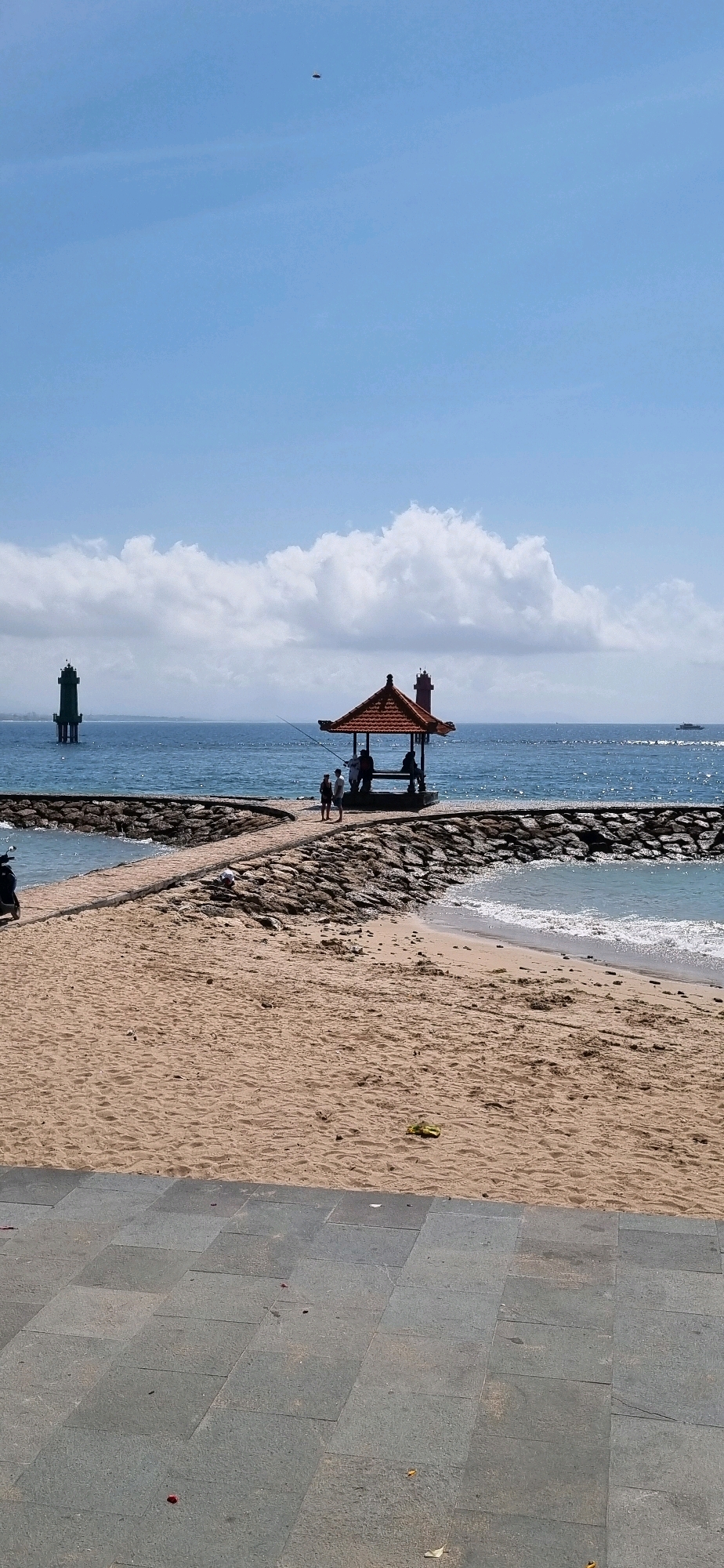 The image captures a serene scene at a beach, likely in Denpasar, Indonesia, given the architectural style of the gazebo and the presence of what appears to be a lighthouse. The foreground is dominated by a sandy beach, with a paved walkway made of large, gray square tiles forming the bottom edge of the frame.
Beyond the beach, a stone jetty extends into the blue ocean, creating a distinct path. Along this jetty, a small, traditional Balinese-style gazebo with a reddish-brown roof stands prominently. Several people are visible near or inside the gazebo, one appearing to be fishing with a long rod.
In the background, the calm blue sea stretches to the horizon, dotted with a few white clouds in the bright sky. Two structures resembling lighthouses are visible in the distance, one on the left, painted green, and another, smaller one to the right of the gazebo, painted red. The overall atmosphere is peaceful and picturesque. The top of the image shows a small, dark, oval-shaped object in the clear blue sky, its nature undetermined.