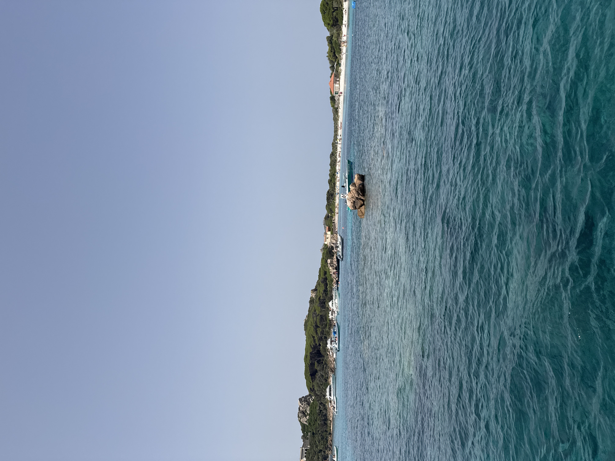 The image captures a picturesque coastal scene with clear blue water and a shoreline dotted with greenery and buildings. The perspective suggests the viewer is on a boat or elevated platform overlooking the water.
In the water, several boats are visible, some anchored near the shore and others further out. One prominent object is a large rock formation near the water's surface, with a small boat positioned beside it. The water itself is a vibrant turquoise, with gentle ripples indicating calm conditions.
Along the coastline, a white sandy beach is lined with lush green trees and vegetation. Buildings, including a structure with a reddish-orange roof, are interspersed along the shore, suggesting a small town or resort. Small groups of people can be seen on the beach, enjoying the sunny weather. The sky is a clear, pale blue, indicative of a bright, sunny day, likely during the day.
The scene evokes a sense of relaxation and vacation, with the beautiful natural landscape and the presence of recreational boats. The location appears to be a popular coastal destination, possibly a bay or cove.
There is no discernible text within the image.