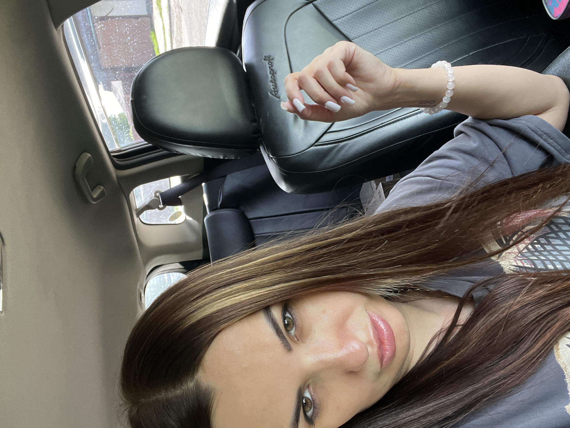A woman is seated in the driver's seat of a car, facing the camera. She has long, dark brown hair with blonde highlights and is looking directly at the viewer with a neutral expression. Her lips are slightly pursed, and she is wearing a grey t-shirt. Her right hand, with neatly painted white nails, is resting on the armrest of the car. She is wearing a white beaded bracelet on her left wrist. The interior of the car, including black leather seats and a seatbelt, is visible. Outside the car window, a brick building and some greenery can be seen. The scene appears to be a sunny day.