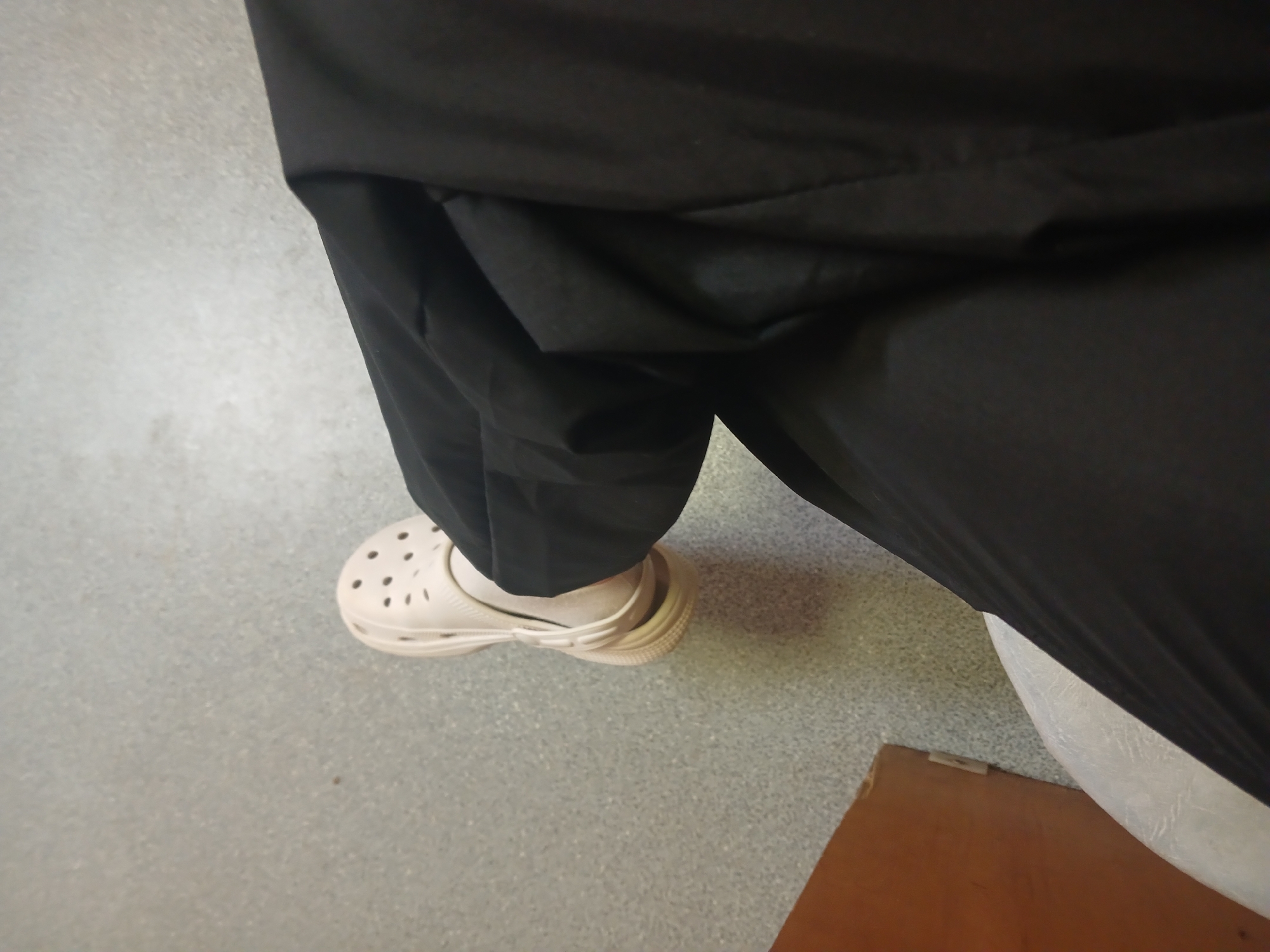 A person is seated, wearing black trousers and beige Crocs-style shoes with white socks. The flooring appears to be linoleum. A section of a wooden surface is visible in the lower right corner, partially obscured by the bottom edge of the seat and a white cushion. The photograph was taken from an overhead perspective.