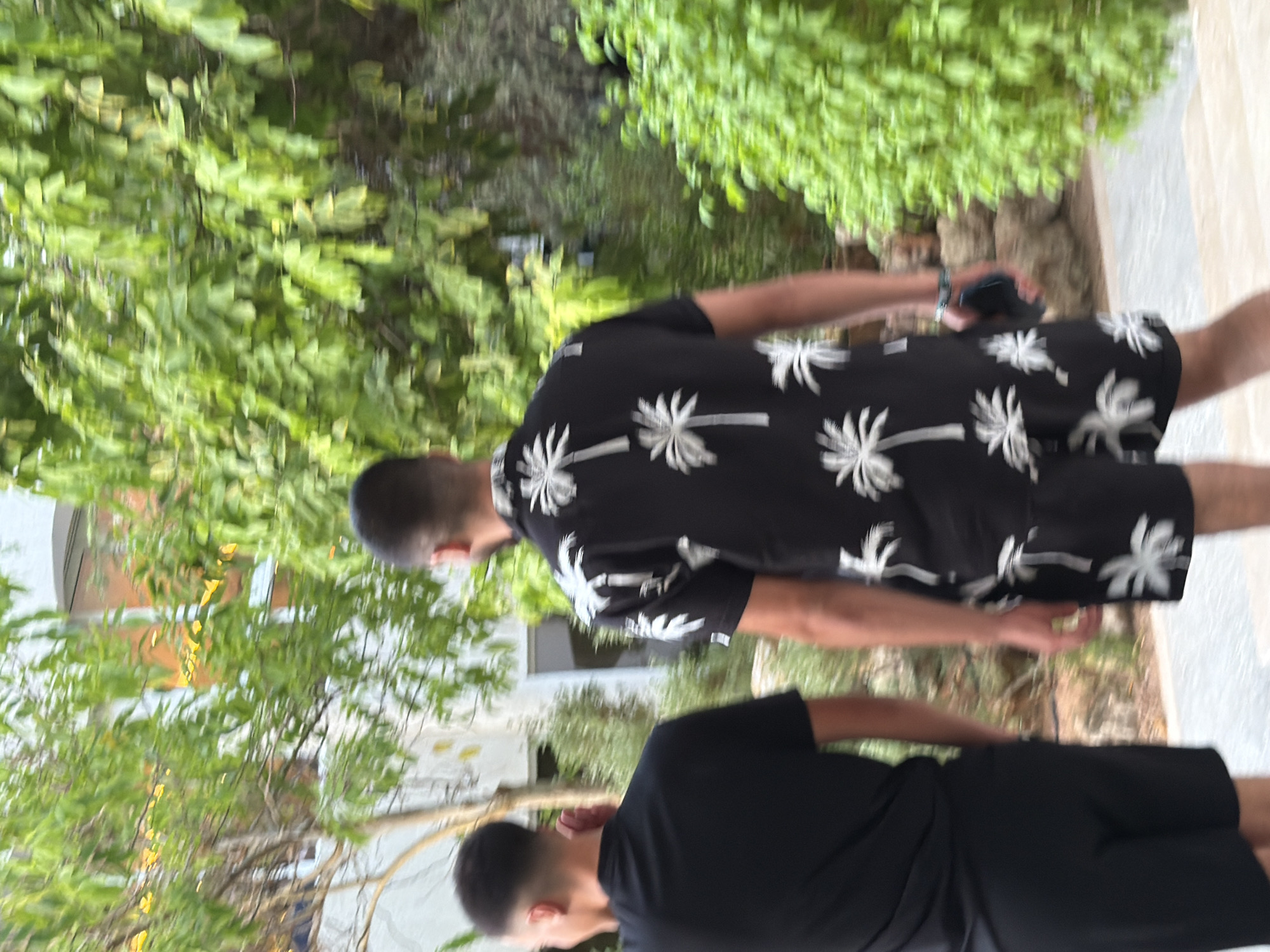 Two individuals, viewed from behind, are walking outdoors amidst lush green foliage, possibly in a resort or garden setting. The person on the right wears a black short-sleeved shirt and shorts adorned with a white palm tree print. This individual is holding a dark rectangular object, likely a smartphone, in their right hand and appears to be wearing a bracelet on their left wrist. The person on the left is dressed in a plain black t-shirt and black shorts. They are walking slightly ahead of and to the left of the first individual. The background consists of dense green leaves from trees and shrubs, with a building visible through the foliage in the upper left quadrant. The lighting suggests it is daytime. The scene is suggestive of a warm climate.