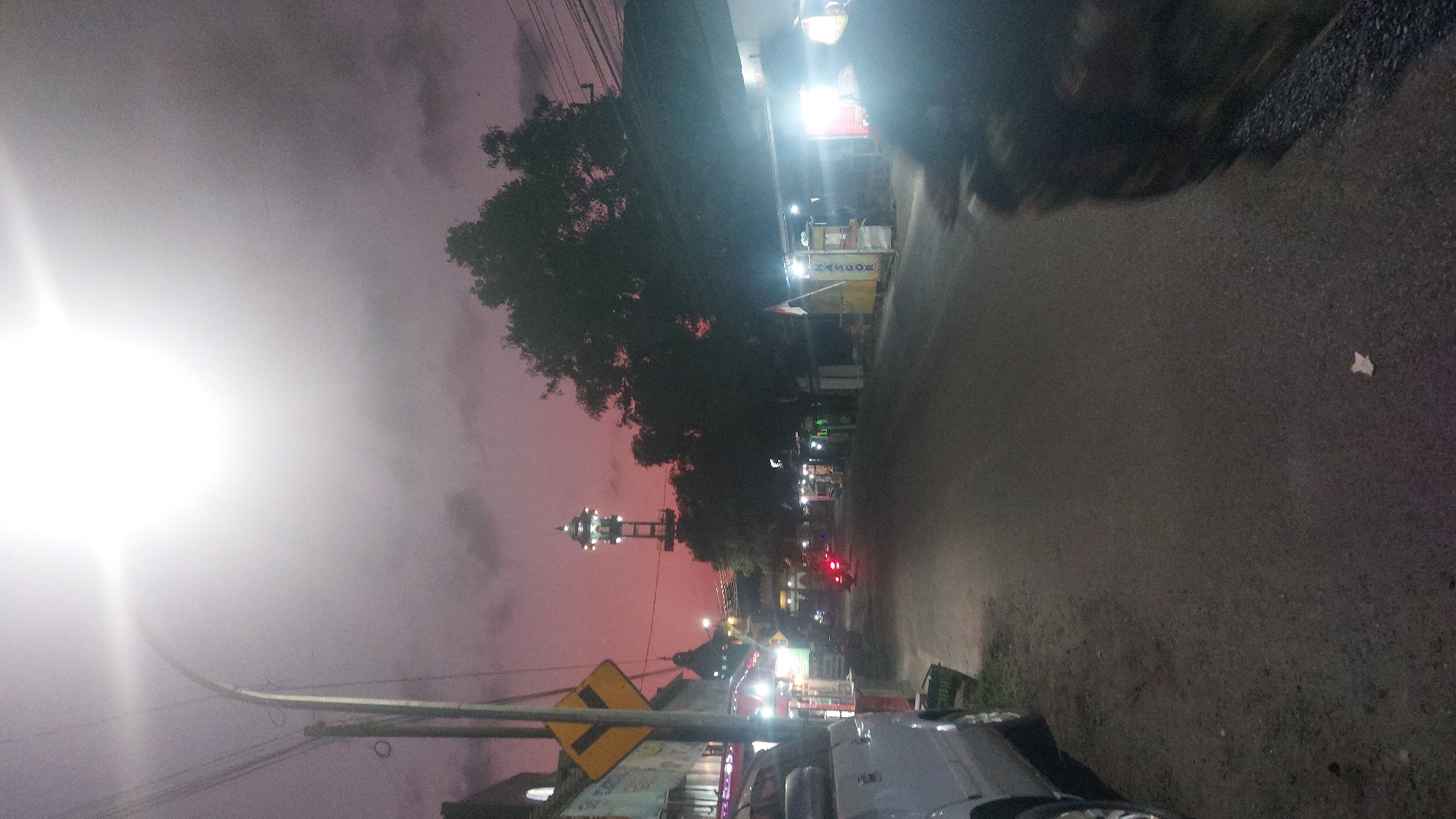 The image depicts a street scene at dusk or night in Sedan, Indonesia, as evidenced by the prominent tower which appears to be a minaret, a common feature of mosques in Indonesia. The sky is a deep purple, suggesting either sunset or twilight, with a bright, overexposed streetlamp dominating the upper portion of the frame. A street curves into the distance, lined with small businesses or stalls illuminated by artificial lights. On the left side of the street, a silver van is parked, partially visible. Further down the street, a minaret with illuminated balconies stands tall against the dusky sky. A silhouette of a person on a motorcycle is visible in the foreground on the right side of the image, their form blurred due to motion. Trees with dark foliage are scattered throughout the scene, adding to the shadowy atmosphere. The overall impression is one of a quiet street in a town in Indonesia during the evening hours.