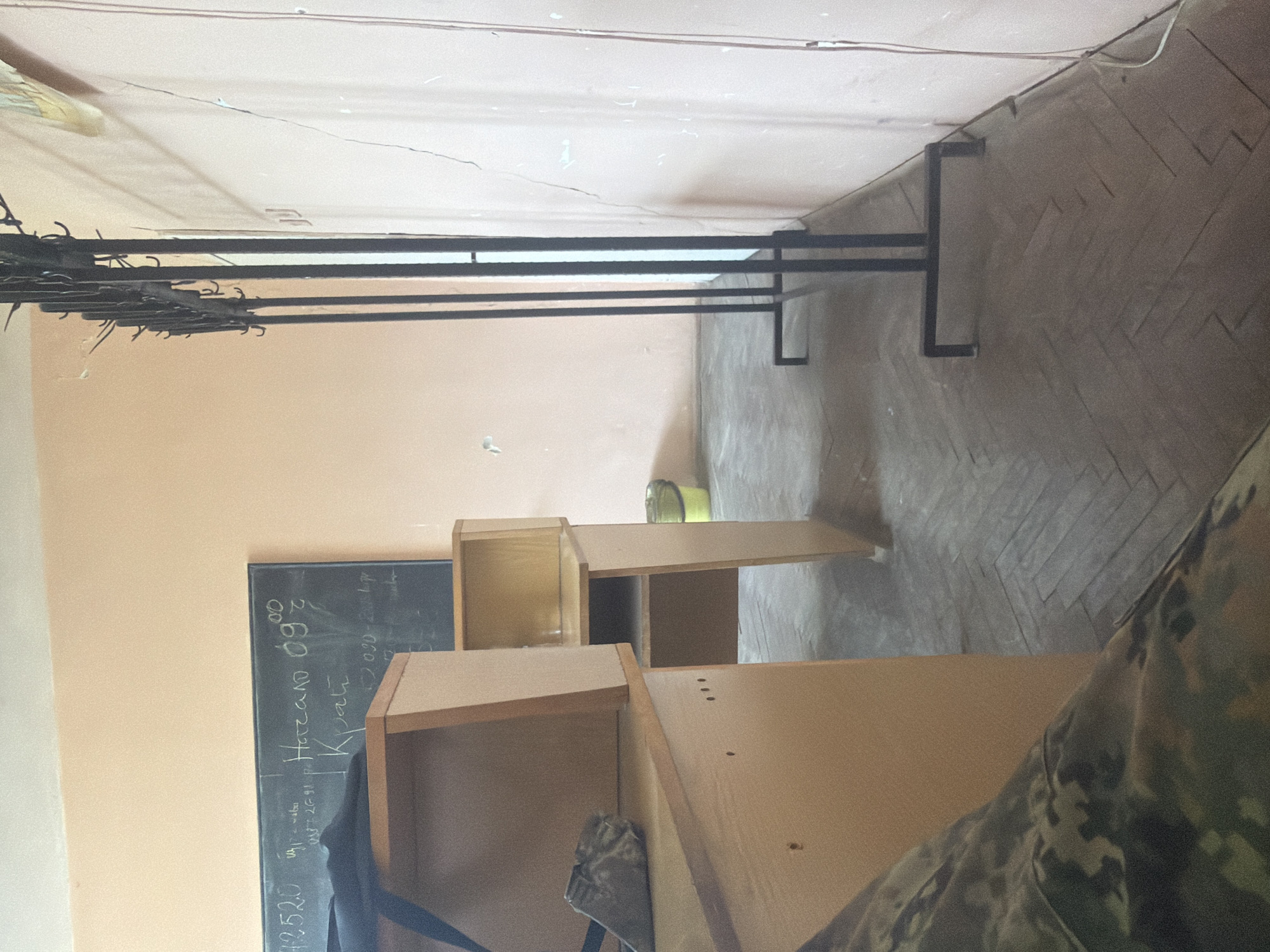 The image shows an interior space in Dolna Mitropolia, Bulgaria. In the foreground, a section of wooden shelving or cabinetry is visible, partially obscuring a blackboard mounted on the wall. The blackboard has handwritten text in a Cyrillic script, potentially containing numerals and short phrases. Above the shelving unit, a metal clothes rack runs along the length of the wall, holding several empty hangers. The floor is covered with patterned tile. A person is present in the photo with a part of the camouflage pattern.
