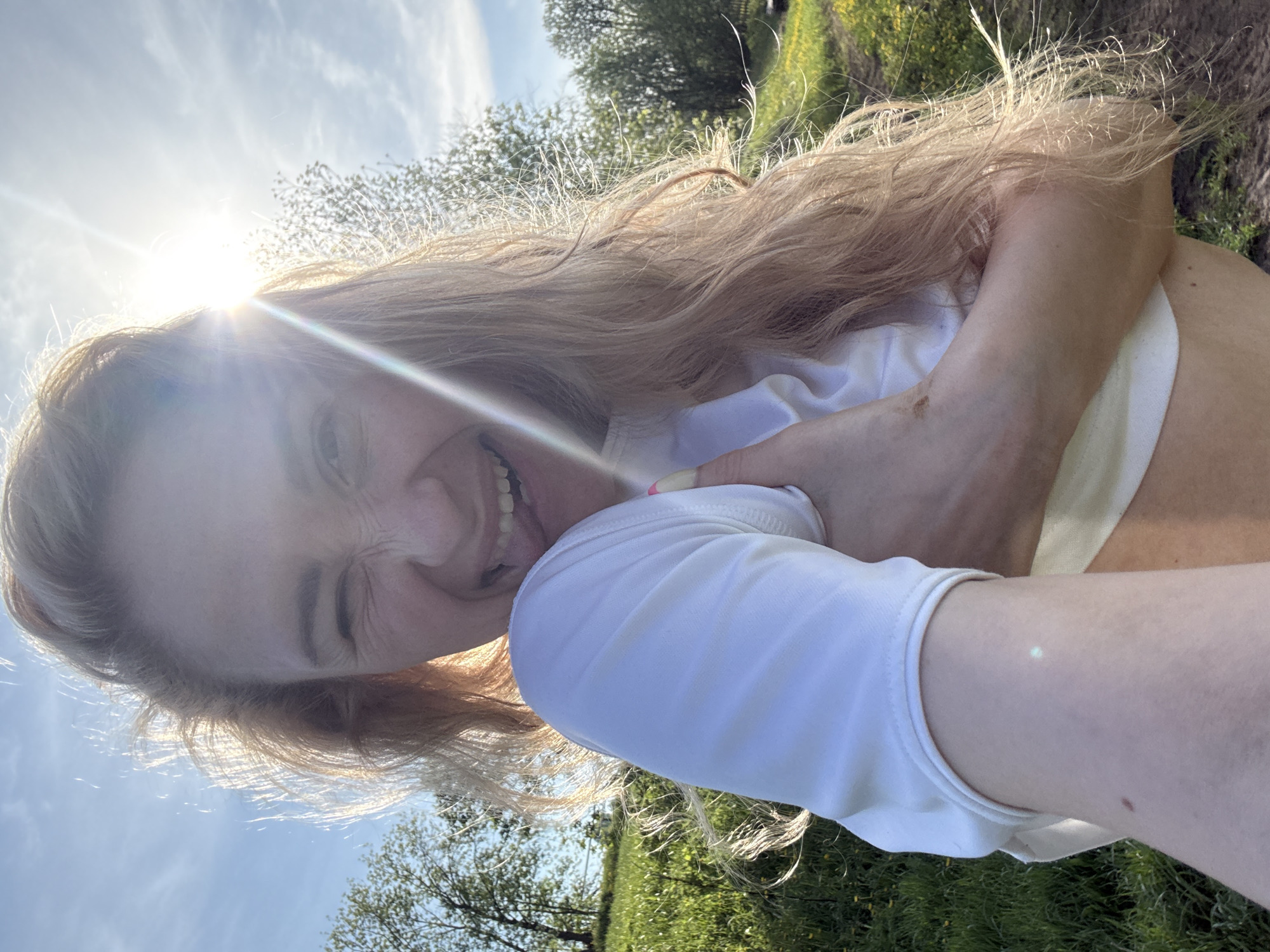 A young woman is taking a selfie outdoors on a sunny day. The photo is taken from a low angle, pointing up towards the woman's face. She has fair skin, blonde hair, and is wearing a white top. Her face is expressive with squinted eyes and a wide smile. The sun is positioned behind her head, causing a lens flare. The background includes a blue sky, trees, and green vegetation. The specific location is not evident, nor is the city of Nizhnyaya Chermoda, Russia.