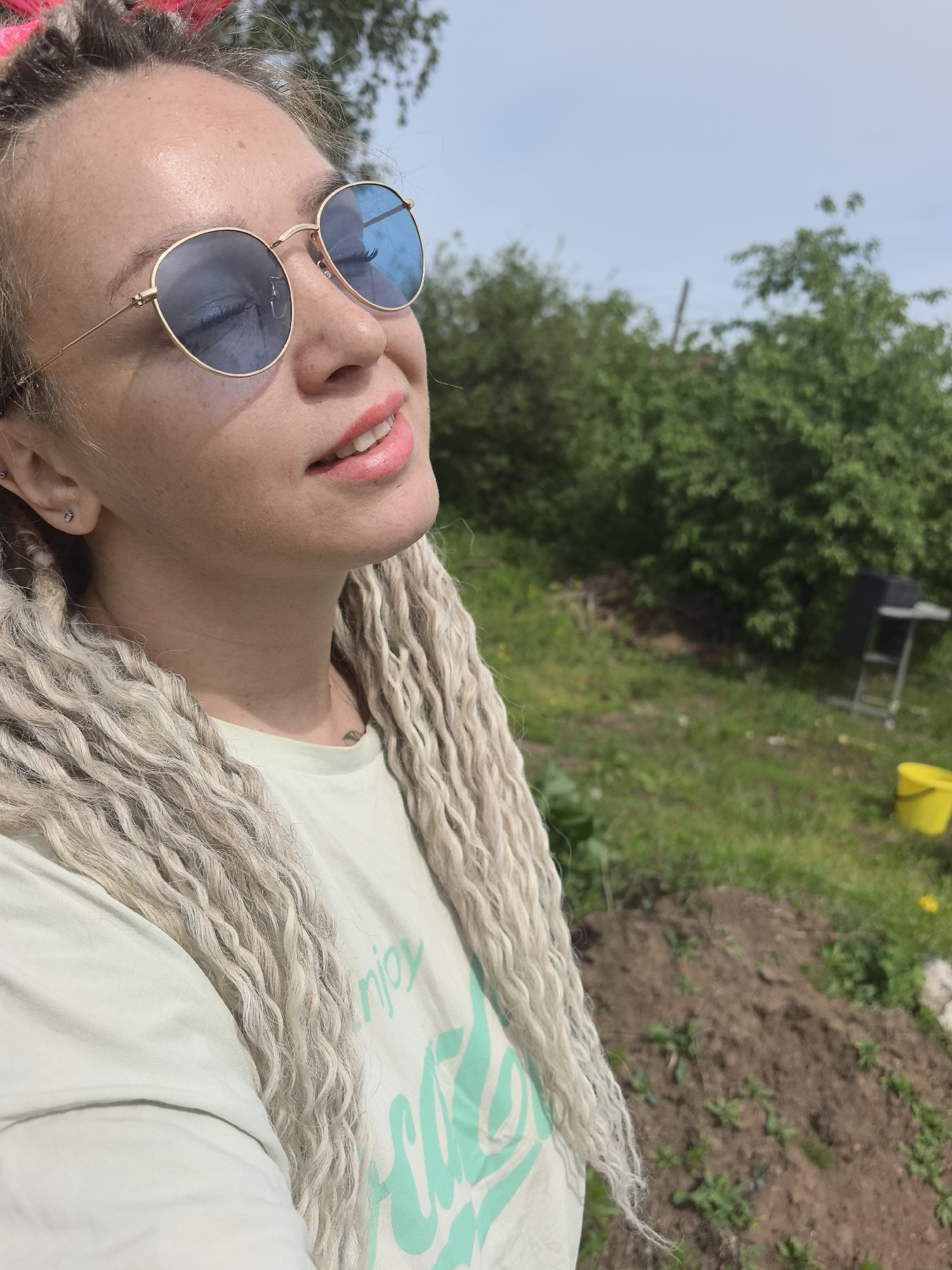 A woman with fair skin and blonde dreadlocks is in an outdoor location, likely a yard or garden. She is wearing round, gold-framed sunglasses with blue lenses and a light green t-shirt with a turquoise design. She is looking up, possibly into the sun. Behind her is a landscape that includes green grass, bushes, a dark chair and table, and a yellow bucket. A pile of dirt is visible in the foreground. The location is the city of Noviy, Russia.