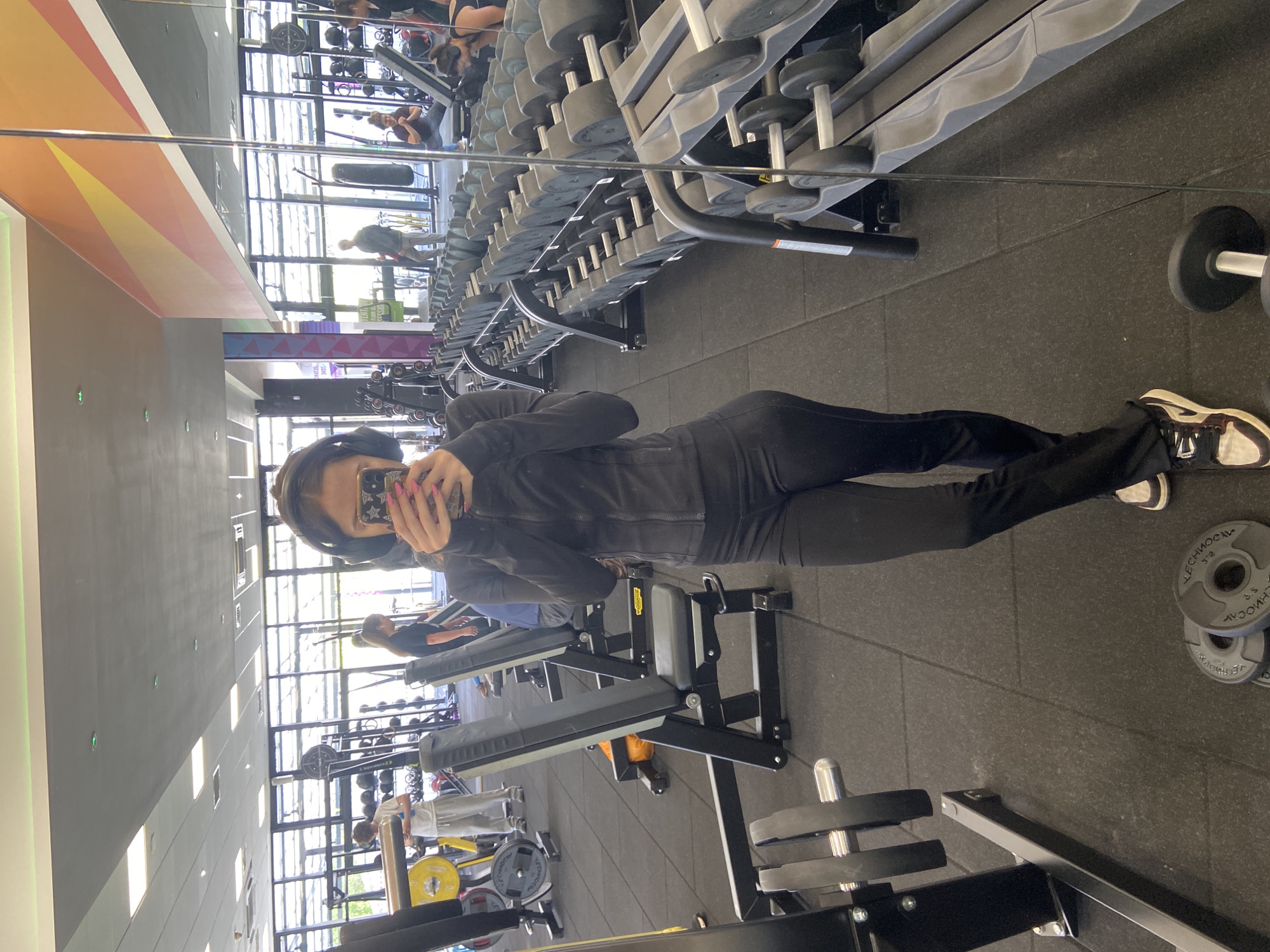 The image appears to be taken in a gym with mirrored walls, reflecting various fitness equipment such as rows of dumbbells, weight machines, and treadmills. In the foreground, a woman is taking a photo of herself in the mirror, wearing dark clothing and headphones. She is holding a mobile phone with a patterned case. The gym has a dark rubber floor and large windows with daylight streaming in. Other people are visible in the background, using the equipment. The location is suggested to be in Mole Valley, United Kingdom.