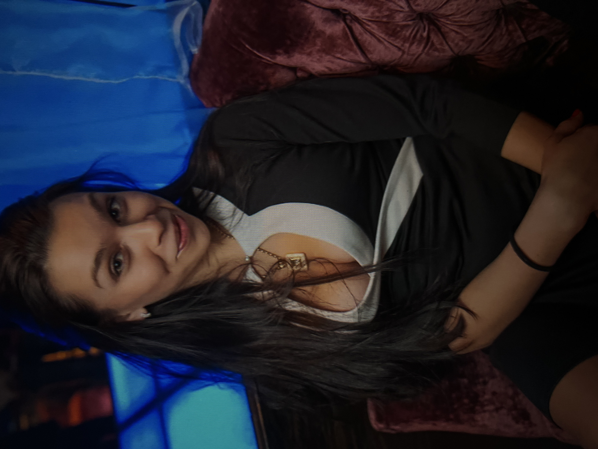 A woman with long, dark hair is seated on a plush, burgundy velvet couch. She is wearing a black and white dress with a plunging neckline and a gold necklace. Her arms are crossed in her lap, and she is looking at the camera with a slight smile. The background is a dark blue, possibly a curtain or wall, with some blurred lights. The scene appears to be indoors, possibly in a nightclub or bar. There are no discernible landmarks or context that would place the image in the city of Qarağandı, Kazakhstan.