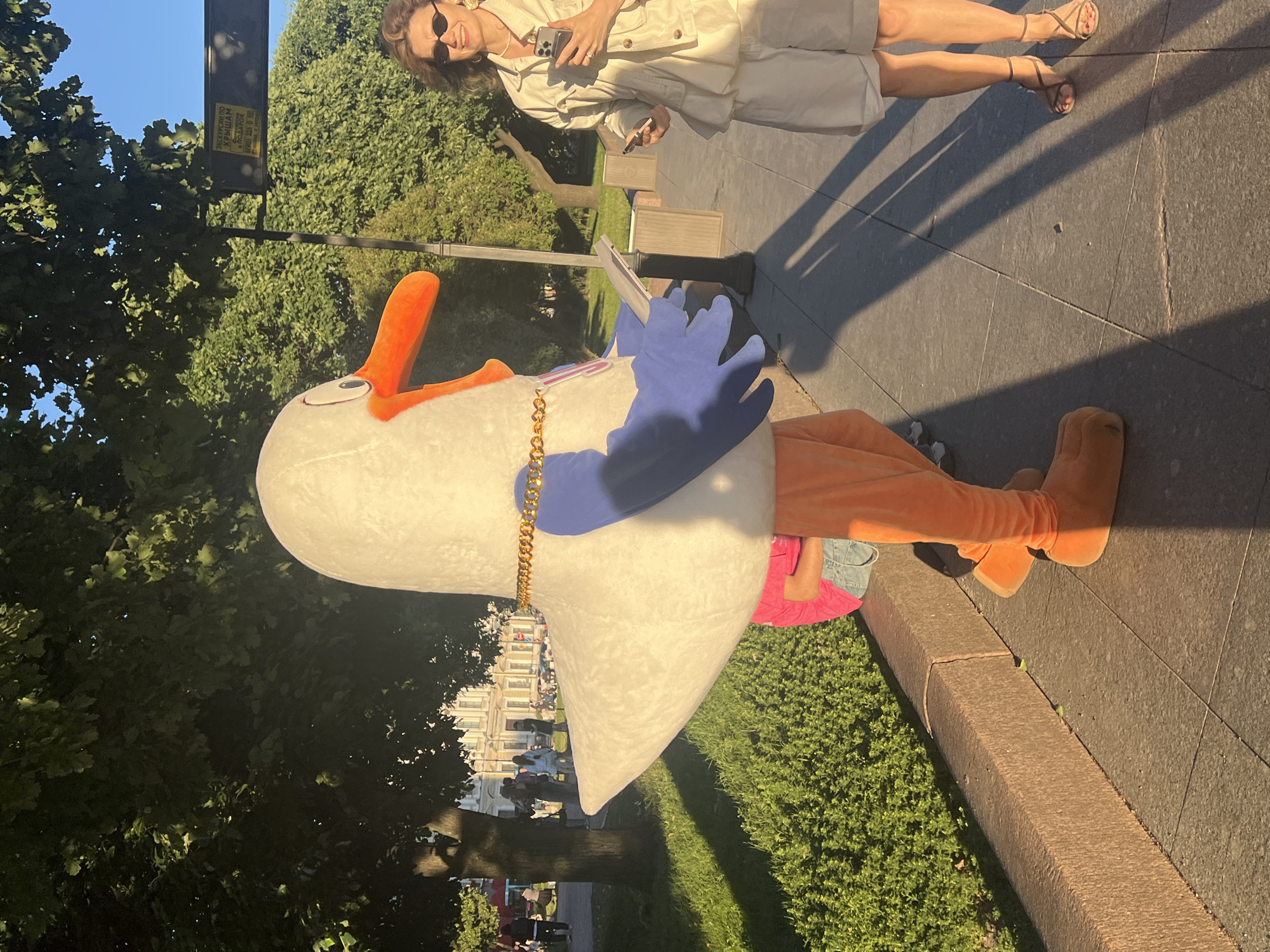 This is a vertical, medium shot photo taken outdoors in what appears to be Saint Petersburg, Russia, during the daytime. The sun is low in the sky, casting long shadows across the pavement, suggesting it's either early morning or late afternoon.
The primary subject is a person in a full-body mascot costume, resembling a white goose with an orange beak and orange legs and feet. The goose costume has a large, round body and is adorned with a gold chain around its neck. The mascot is holding a tablet or book in its blue-gloved hands.
In the background, a woman wearing sunglasses, a light-colored jacket, and shorts is visible, holding a smartphone. Her expression appears happy and relaxed. The setting includes a paved walkway with grassy areas and trees. Behind the woman, there is a lamppost with a sign attached, though the text on the sign is mostly illegible due to its distance and the angle of the photo. Further in the background, a light-colored, ornate building, possibly a historical structure, is partially visible through the trees. The overall mood of the photo is cheerful and relaxed, likely capturing a moment of informal street performance or promotion.