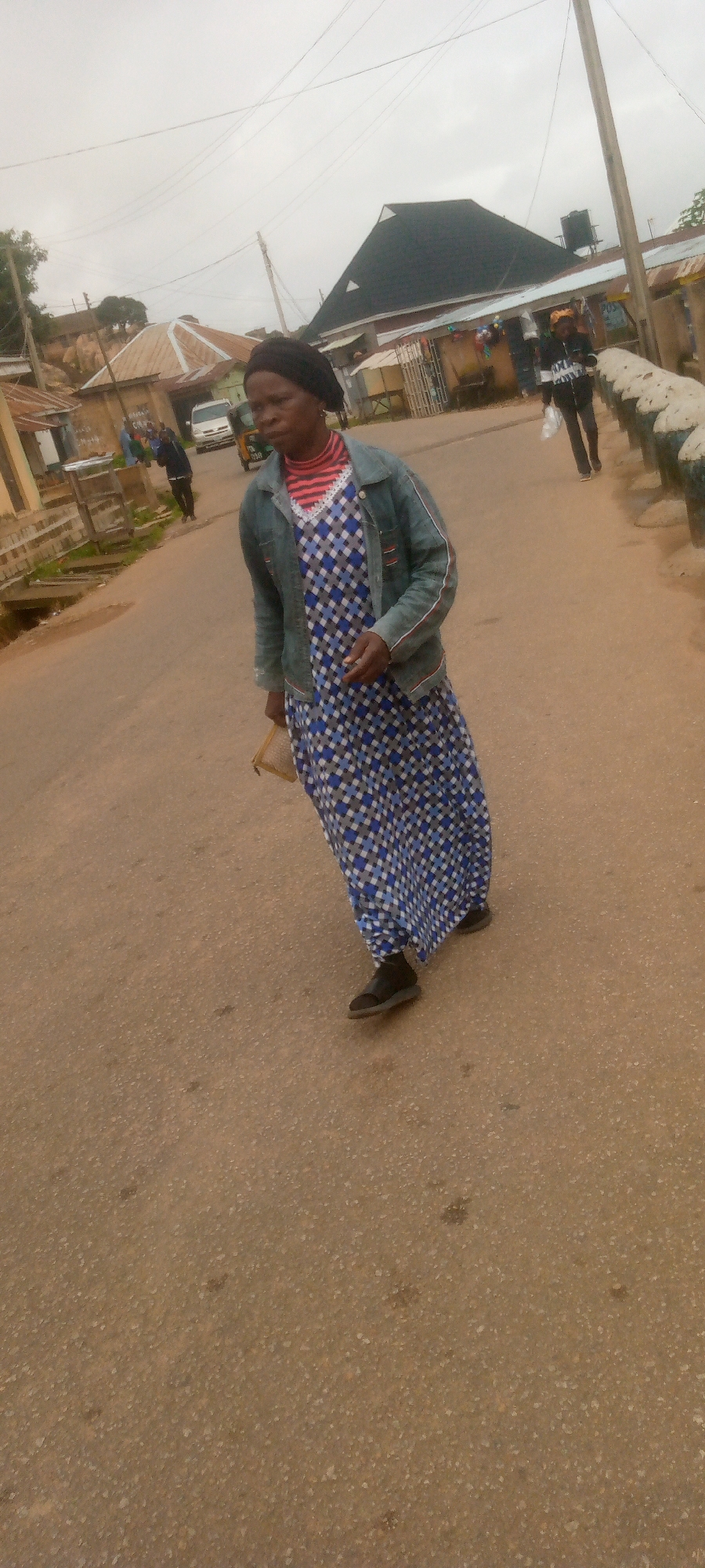 A medium shot captures a Black woman walking on a paved road in what appears to be a town or city. She is wearing a dark blue denim jacket over a white, blue, and grey checkered dress. Underneath the dress, a red and white striped shirt is visible at the neckline. She has a dark headscarf covering her hair and is carrying a light-brown rectangular clutch in her right hand. Her feet are clad in black sandals.
The road surface is made of asphalt, appearing worn and somewhat uneven. In the background, to the left, are several low-rise buildings with corrugated metal roofs, some exhibiting signs of wear. Further back, a white vehicle and a three-wheeled auto-rickshaw are visible. To the right of the road, a low concrete barrier with decorative elements runs along the sidewalk. Behind this barrier, more buildings are visible, including one with a dark, multi-faceted roof structure. The sky is overcast, with a pale grey cloud cover. Power lines crisscross the sky. The overall lighting suggests an overcast day. The location is identified as Unknown, Unknown.