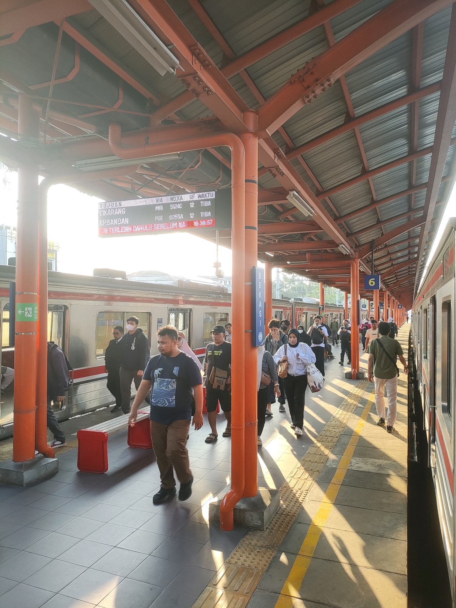 The image depicts a train station platform in Bekasi, Indonesia, bathed in the warm light of sunrise or sunset. A train is parked alongside the platform, its silver and red exterior visible. A digital display board above the platform shows train schedule information, including destinations like CIKARANG and KP.BANDAN.
Several people are present on the platform, some boarding or disembarking the train, while others are waiting. They are dressed in casual attire, and a few are wearing face masks. Among the visible objects are red suitcases, benches, and support pillars painted orange. The platform itself is paved with grey tiles, marked with yellow lines and tactile paving for the visually impaired. Overhead, a corrugated metal roof supported by orange metal beams covers the platform, providing shelter. The environment appears to be an active transit hub, with the sun casting long shadows across the platform.