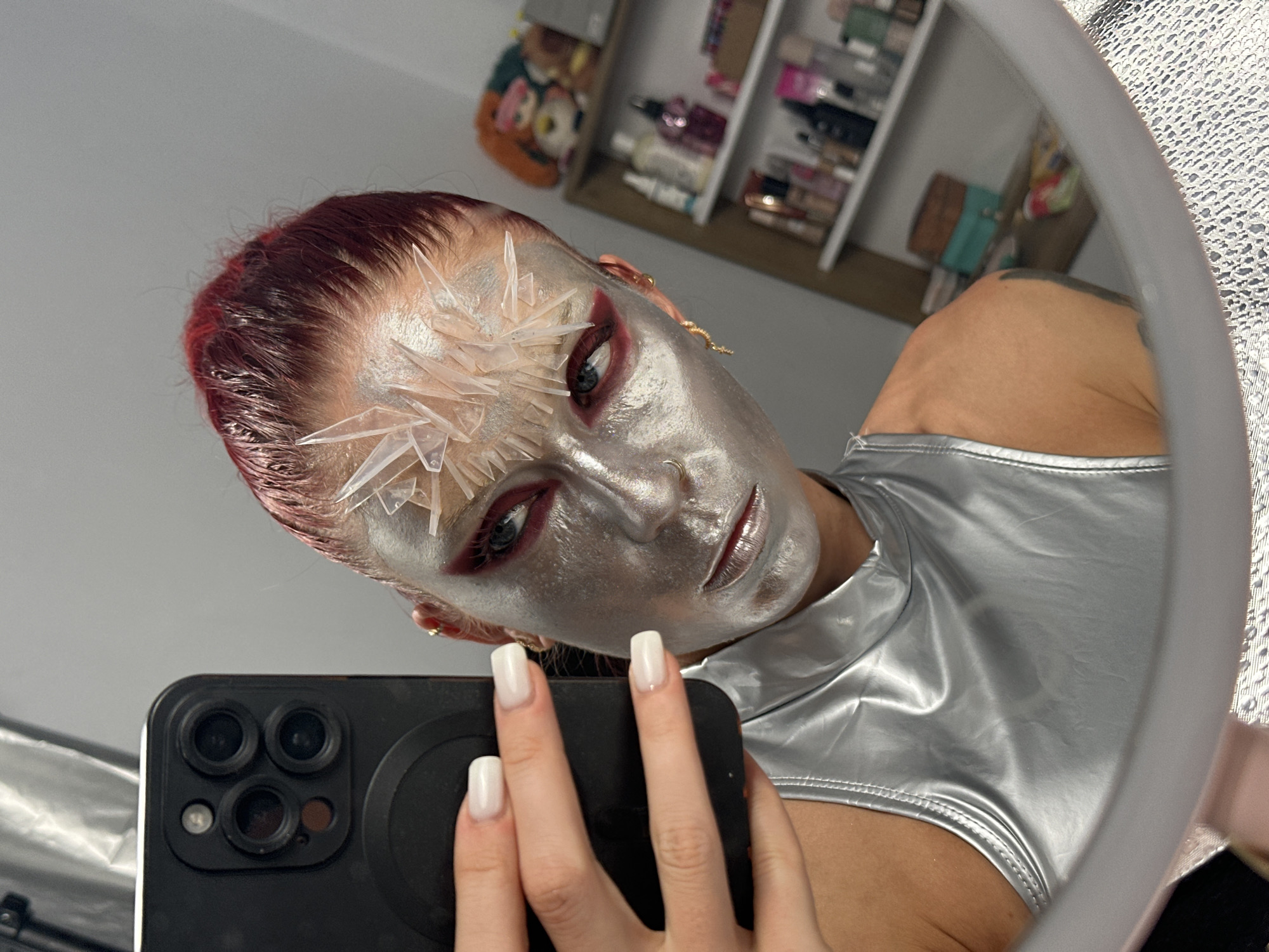 A person is taking a selfie in a mirror. They have applied metallic silver body paint and some crystal-like accessories to their face, with dramatic red eyeshadow. Their hair is pulled back and dyed a reddish-purple color. They are wearing a shiny, silver, sleeveless top. In the background, a shelf is visible with various bottles and containers, possibly cosmetics or toiletries. The image is taken indoors, likely in a personal space. The location is not specified in the image.