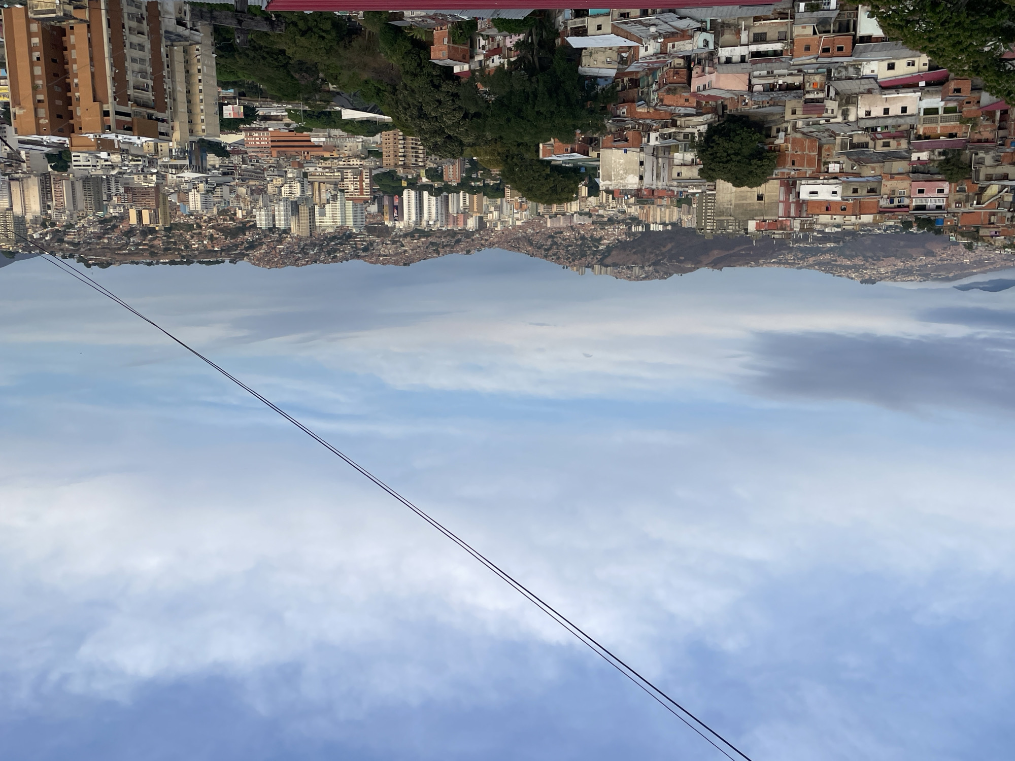 The image shows an upside-down view of Caracas, Venezuela. In the foreground, there is a blue sky with clouds and a power line. In the middle ground, there is a city with a mix of high-rise buildings, houses, and favelas. In the background, there are mountains.