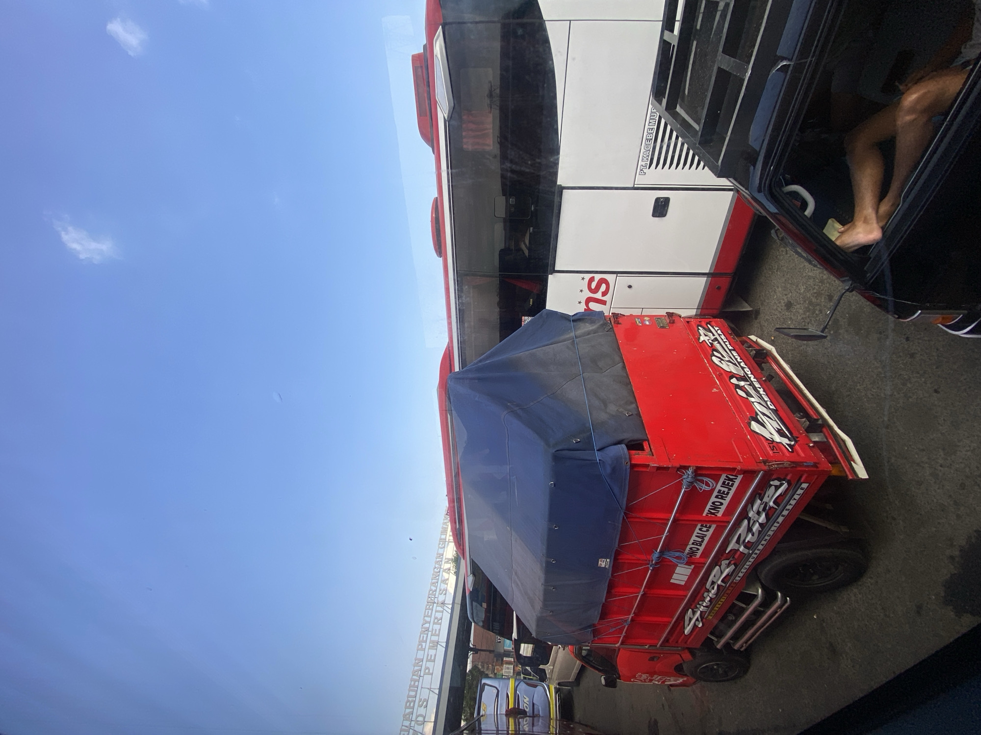 In Melaya, Indonesia, under a clear blue sky, a traffic jam is captured. A red truck with a blue tarp covers its cargo in the foreground, parked beside a white and red bus. Text on the bus reads "PT. KAGEBE MUD". To the right, a black vehicle has its door open, revealing a person's legs stretched out inside. A sign overhead reads "ABUHAN PENYEBRANGAN GILIMANUK". The scene depicts a typical day at a transportation hub with various vehicles waiting.