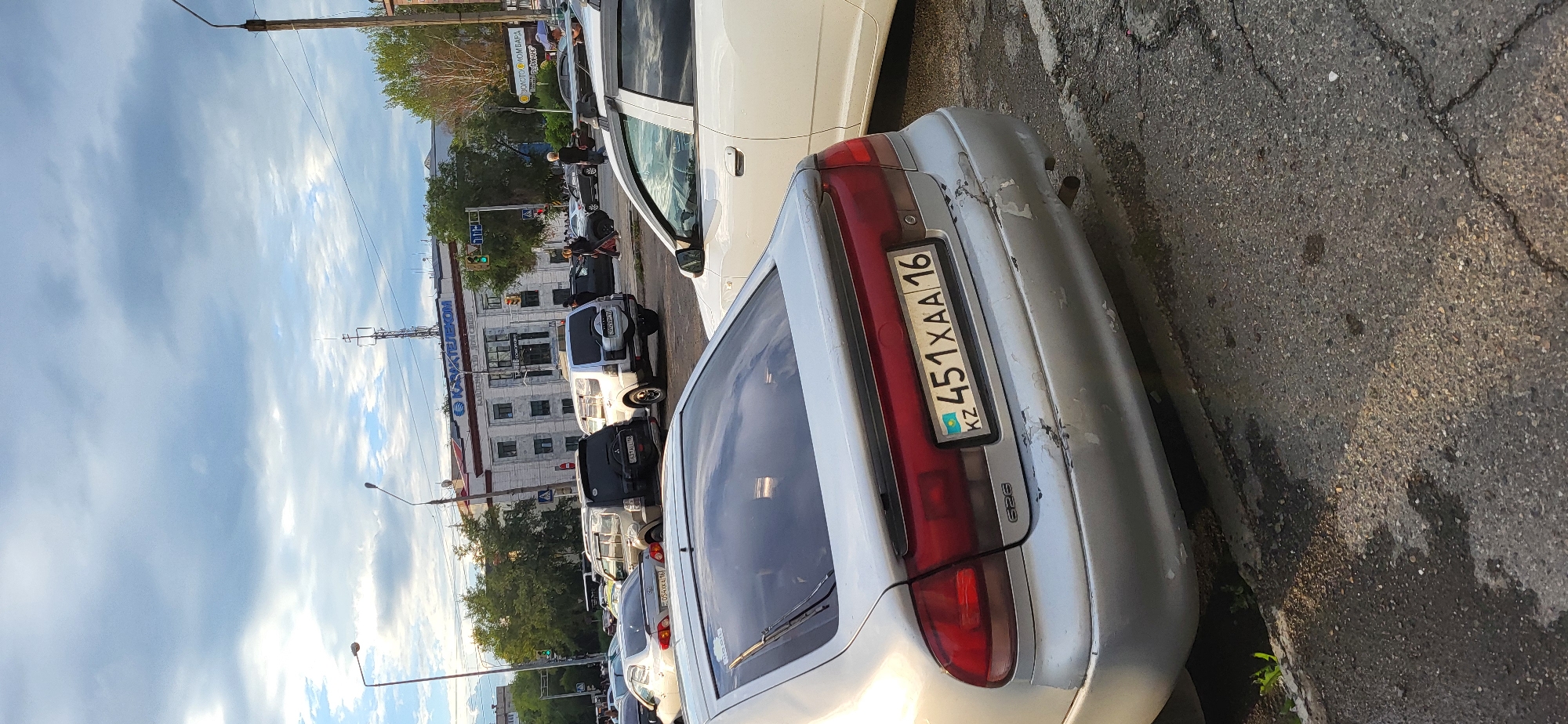 The image captures a street scene in the city of Oskemen, Kazakhstan, under an overcast sky. In the foreground, a silver Mazda 626 is parked, its rear bumper showing significant damage with peeling paint. The license plate of this car reads "KZ 451 XAA 16". To the right of the Mazda, the rear of a white sedan is visible. In the middle ground, several other vehicles are parked, including a black SUV and two silver SUVs, one of which has a license plate starting with "KZ". Beyond the vehicles, a multi-story building with "КАЗХТЕЛЕКОМ" signage is prominent. The surrounding environment includes trees, streetlights, traffic signals, and pedestrian walkways, indicating a commercial or mixed-use urban area. Several individuals are walking in the background, suggesting daily activity.