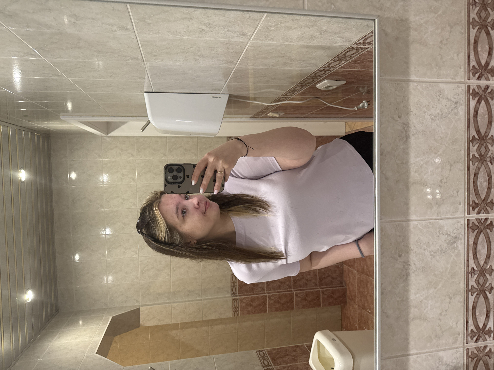 A woman is taking a selfie in a bathroom. The bathroom has beige and brown tiles. A hand dryer is mounted on the wall above a toilet. The location is likely a public restroom in the city of Chaikovsky, Russia. The woman is wearing a white T-shirt and black pants. She is holding a smartphone in her right hand. She has blonde and brown hair and is wearing sunglasses on her head.