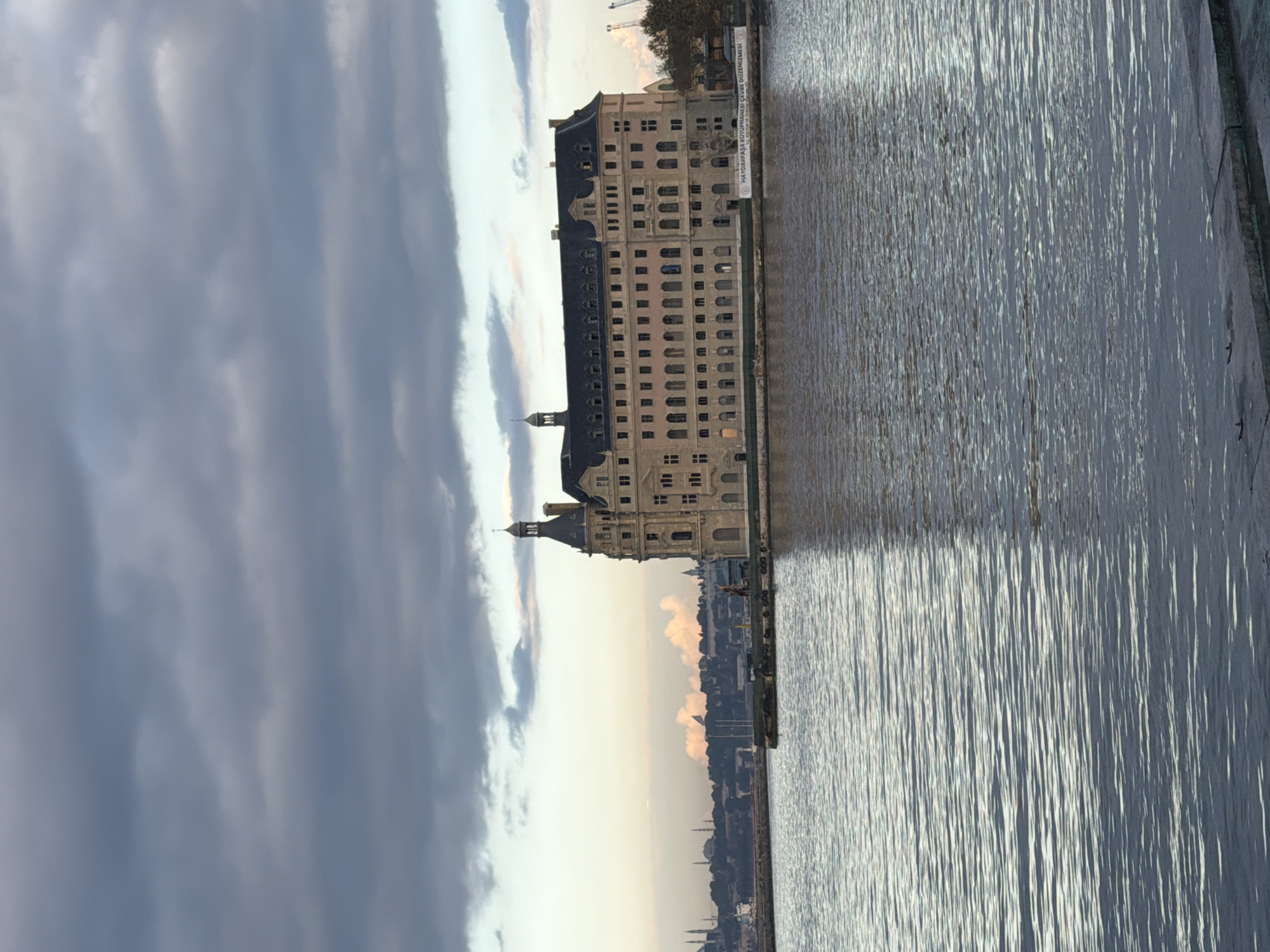 The image shows the Haydarpaşa Train Station, a historic building located on the Bosphorus Strait in the Kadıköy district of Istanbul, Turkey. The station is a large, imposing structure with a distinctive clock tower and a long, multi-story facade. It is made of stone and has numerous windows. The building is situated on the waterfront, with the calm waters of the strait in the foreground.
The sky above is partly cloudy, with dramatic, dark clouds contrasting with patches of lighter sky. The water reflects the sky and the building, creating a shimmering effect. In the distance, the Istanbul skyline can be seen, with minarets and other buildings dotting the horizon. There are also some cranes visible in the background, suggesting ongoing construction or industrial activity.
A banner with Turkish text is visible on the side of the building, near the water's edge. The overall impression is one of a grand historical landmark set against a backdrop of a dynamic urban landscape and moody weather.