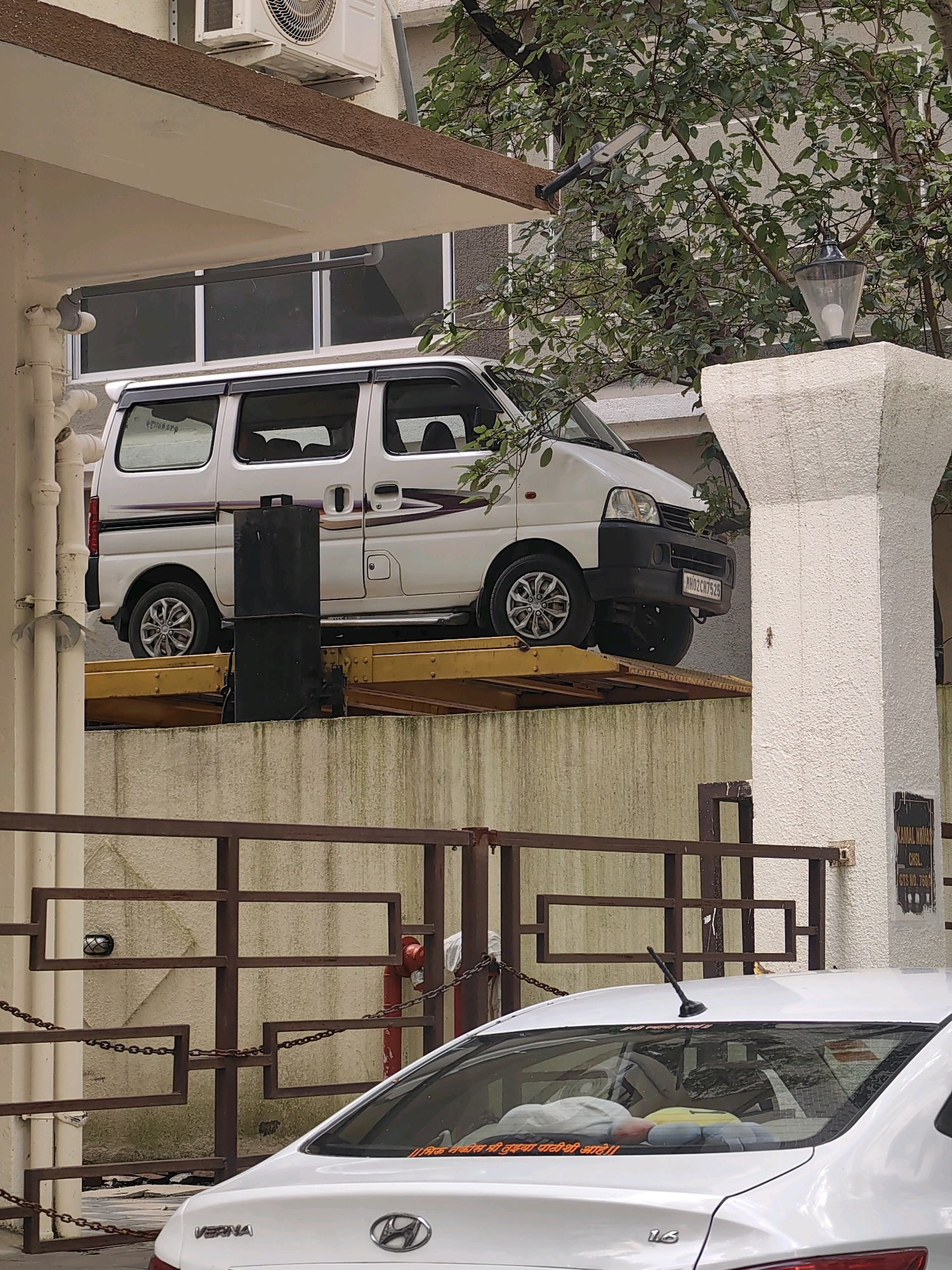 In Mumbai, India, a white van is parked on a yellow mechanical car lift on a raised platform. The van has a license plate that reads "MH02CH7525". In the foreground, the rear of a white Hyundai Verna car is visible, with a sticker on its rear windshield. The environment includes a building with an air conditioner unit, a lamp post, trees, and a metal gate with chains. The overall scene appears to be an outdoor parking area or driveway.