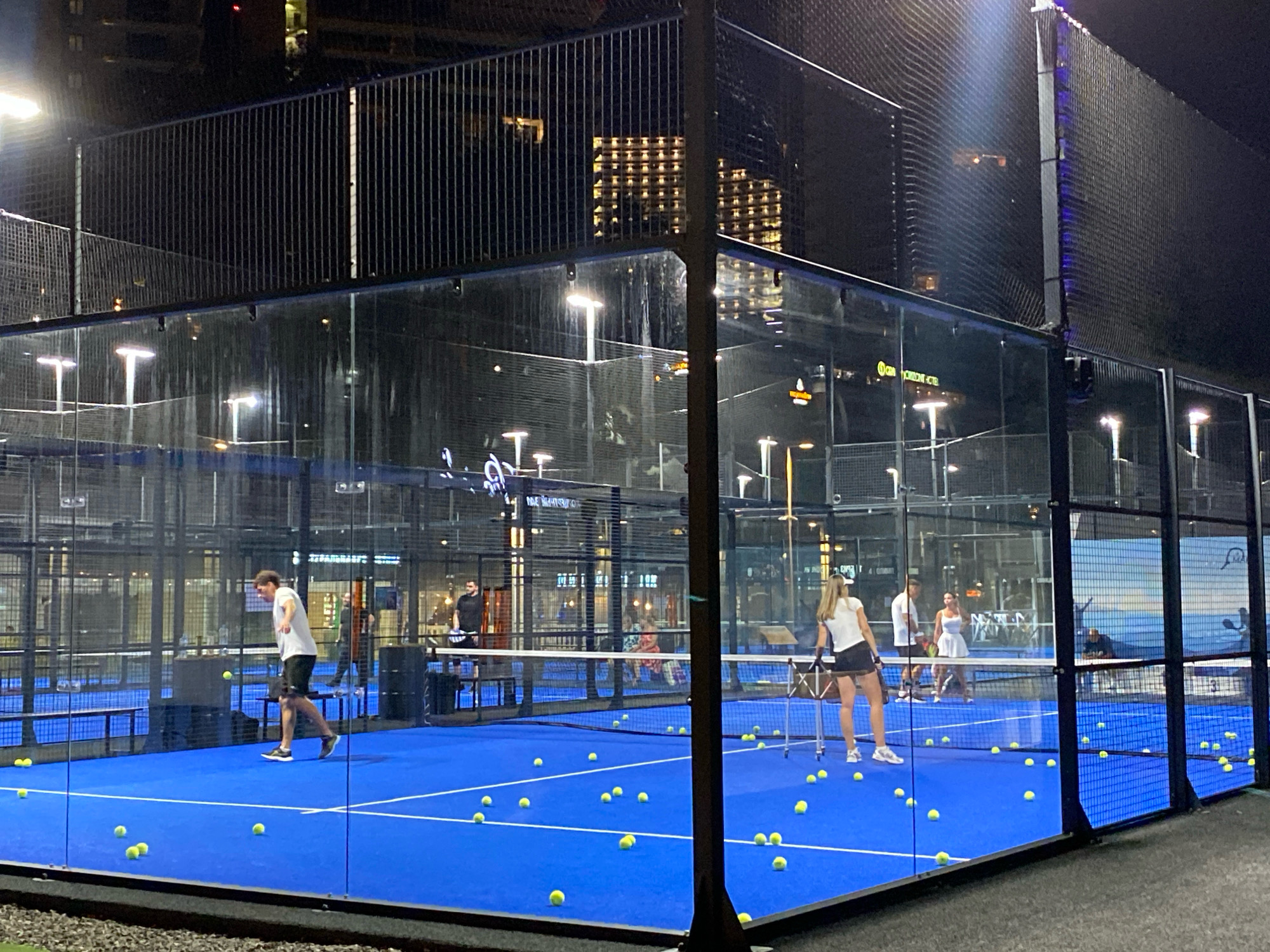 The image depicts an outdoor padel tennis court at night in Sochi, Russia. The court has a bright blue surface with white lines marking its boundaries. The court is enclosed by glass walls and a metal fence. Numerous yellow tennis balls are scattered across the court. Several people are visible playing or watching the game. In the foreground, a man in a white t-shirt and black shorts is positioned on the court. Further back, a woman in a white top and black skirt is visible, as are other individuals in the background. The surrounding area is dark, illuminated by artificial lights on the court and lights from buildings in the distance. The urban environment of Sochi is suggested by the background cityscape.
