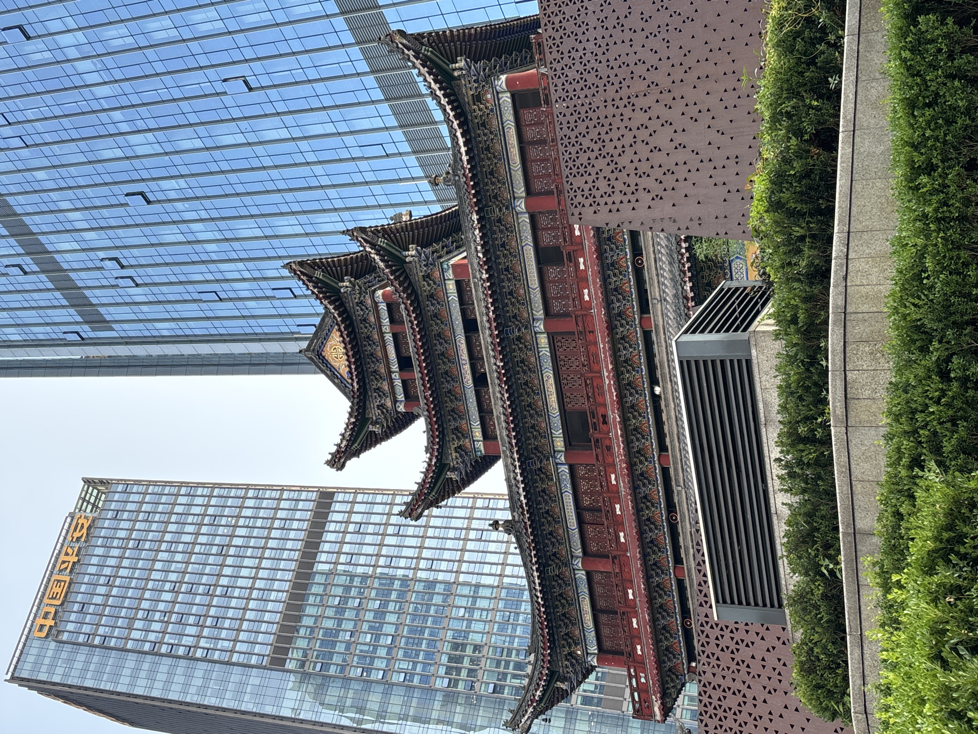 This is a vertical, medium-shot photograph taken in the Yuzhong District of China, featuring a juxtaposition of modern and traditional architecture. The foreground is dominated by the ornate, multi-tiered roof of a traditional Chinese building, painted in vibrant reds and blues with intricate patterns and carvings. These roofs curve upwards at the eaves, characteristic of ancient Chinese design.
Behind this traditional structure, two modern skyscrapers rise. The one on the left is a tall, glass-faced building with a grid-like pattern of windows, reflecting the sky. The skyscraper on the right is partially obscured by the traditional building and greenery but also appears to be a modern office building with a patterned facade.
To the far right, dense green foliage forms a natural wall, contrasting with the man-made structures. A textured, dark brown wall with a repeating triangular pattern is visible between the traditional building and the greenery.
The sky is a clear, bright blue, suggesting a sunny day. The lighting indicates it is likely daytime, possibly late morning or early afternoon. There is no visible activity of people.
Visible text includes golden Chinese characters on the glass skyscraper on the left, which read "中国平安" (China Ping An). This text is a key location cue, identifying a specific building or company. The scene captures a blend of old and new, reflecting urban development where historical architectural styles are preserved or integrated alongside contemporary construction.