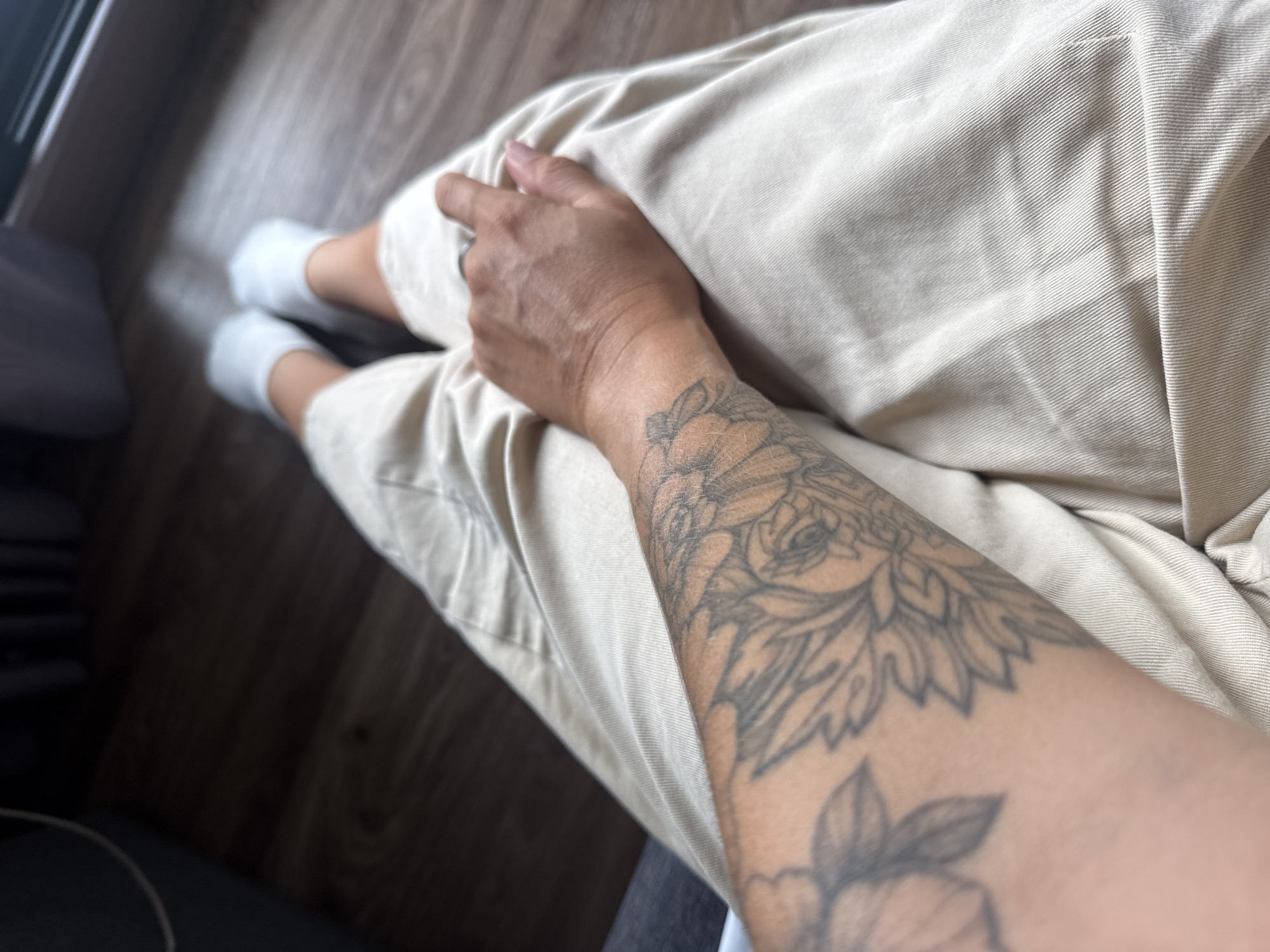 A person's legs, arms, and hands are visible, wearing light-colored pants and white socks. The person has a tattoo on their left forearm featuring floral designs. Their right hand rests on their lap, wearing a ring on the index finger. The background appears to be a wooden floor, suggesting an indoor setting. There is no identifiable information to place this scene in Bangkok, Thailand.