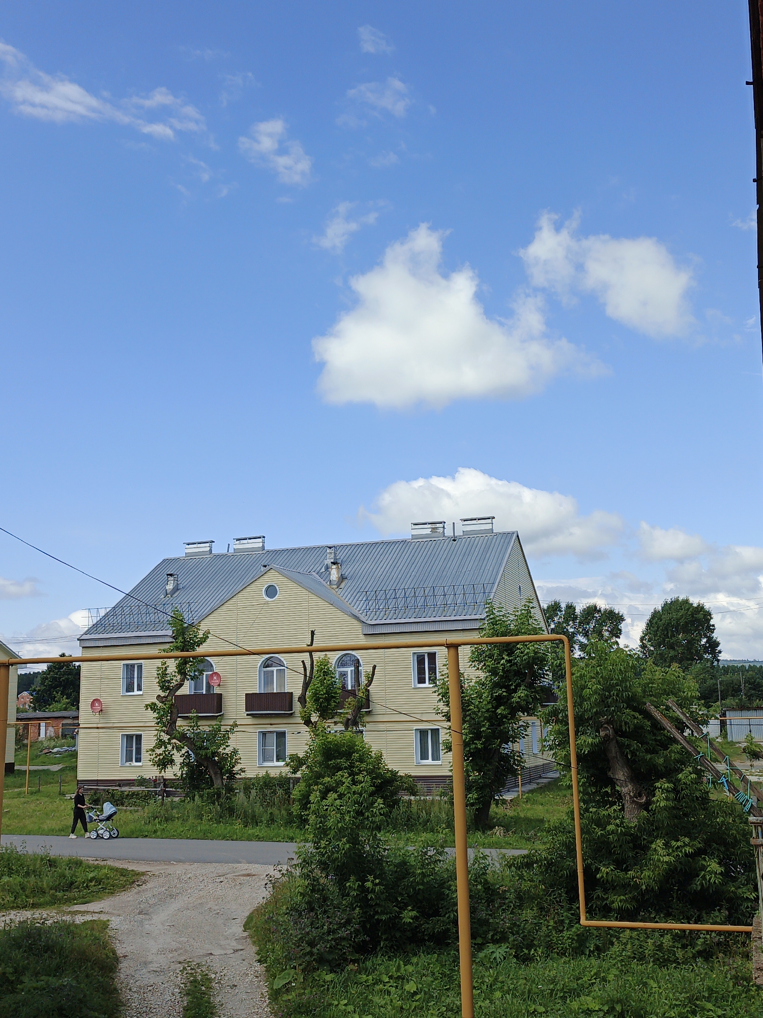 In Medvedyovka, Russia, a pale yellow, two-story building with a gray metal roof stands under a bright blue sky with scattered white clouds. The building features multiple windows, some arched, and satellite dishes mounted on the exterior. A woman walks a stroller along a paved road in front of the building, which is surrounded by lush green trees and vegetation. A gravel path leads away from the road towards the foreground. Yellow pipes, likely part of a gas line, are visible in the lower right portion of the image. The scene appears to be a residential area in a rural or semi-rural setting.