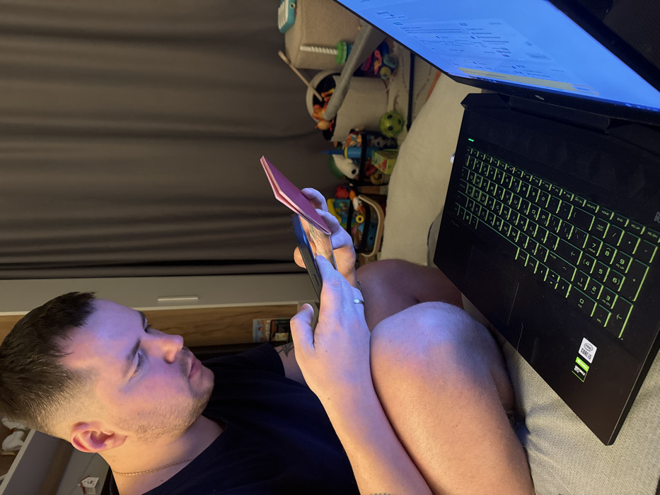 A man lies on a couch, holding a phone and a pink card in his hands, seemingly taking a picture. A laptop sits on his legs, its screen lit up and keyboard illuminated with green lights. Behind him, a grey curtain hangs. A shelf containing toys and other miscellaneous items is also visible in the background.