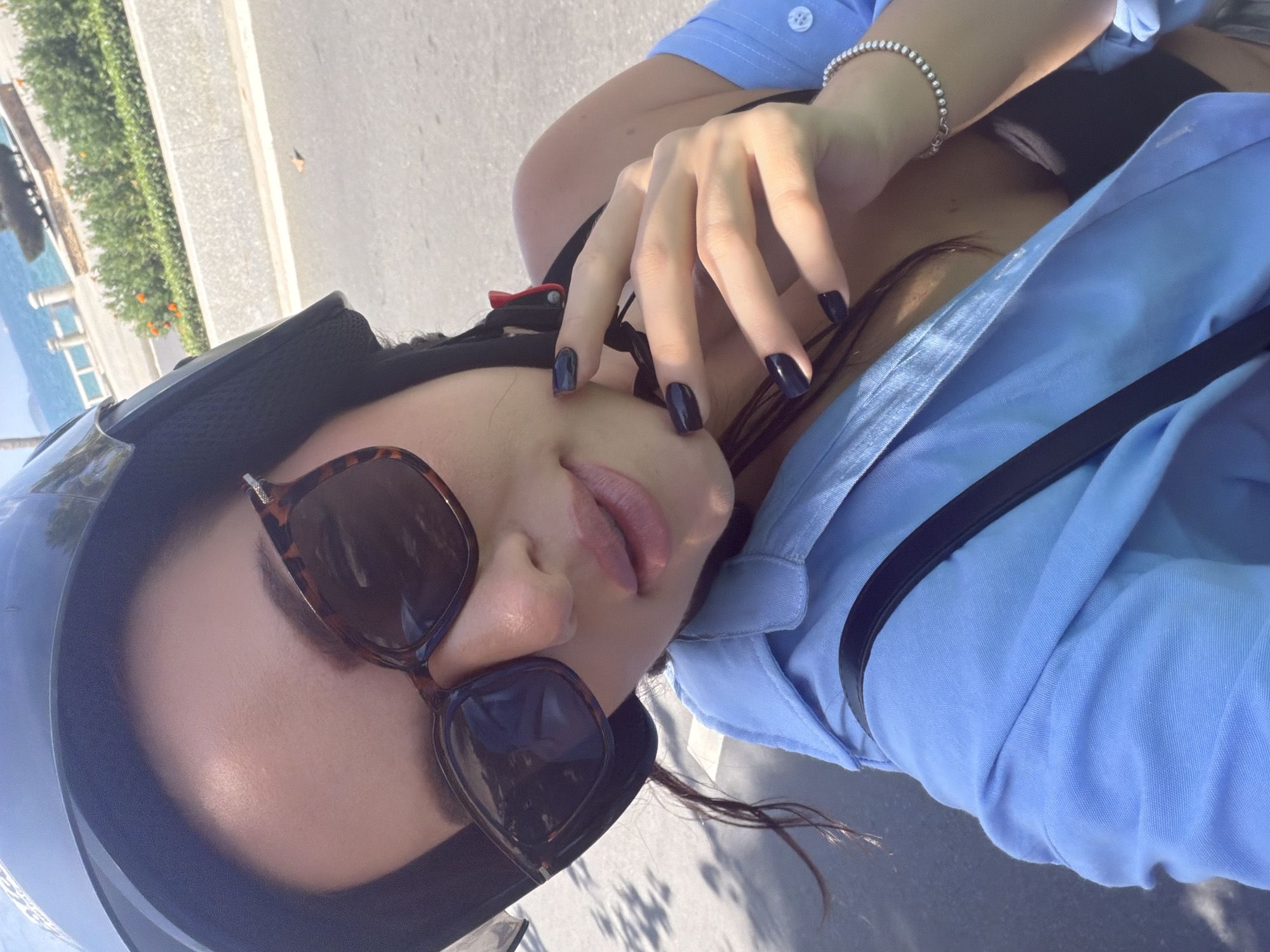 A woman is depicted from a close-up perspective, wearing a dark helmet, tortoiseshell-patterned sunglasses, and a light blue unbuttoned shirt over a black undergarment. Her dark hair is partially visible, and her fingernails are coated with dark, glossy polish. A silver beaded bracelet is present on her left wrist, and a black strap from a bag crosses her shoulder. Her left hand is raised to her jawline. The background reveals an asphalt road to the left, and to the right, a coastal environment identified as Nha Trang Ward, Vietnam. This area includes a concrete curb, green shrubbery containing orange blossoms, a white railing, and the expanse of the blue ocean under a brightly lit sky. A dark-toned monument is partially visible beyond the greenery.