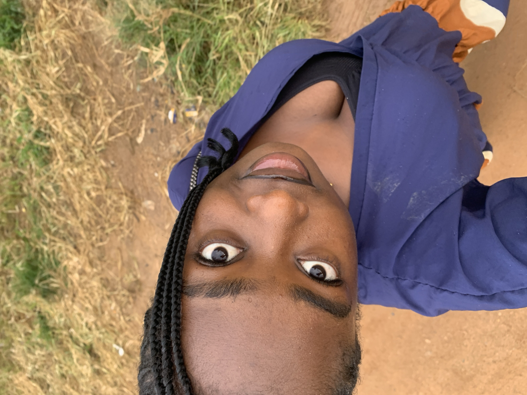 A close-up, overhead shot shows the face of a young Black woman. She has dark brown skin, braided black hair, and appears to be wearing makeup. Her eyes are wide open, and her mouth is slightly agape. She is wearing a blue jacket over a black top, and what appears to be orange and white patterned shorts. The background consists of dry grass and dirt. The image is taken from a high angle, suggesting the photographer was standing above the subject. The photo is oriented such that the woman's head is at the bottom of the frame, and her feet are towards the top. The image is outdoors, possibly in a rural setting or a less developed urban area, given the presence of dry vegetation and dirt.