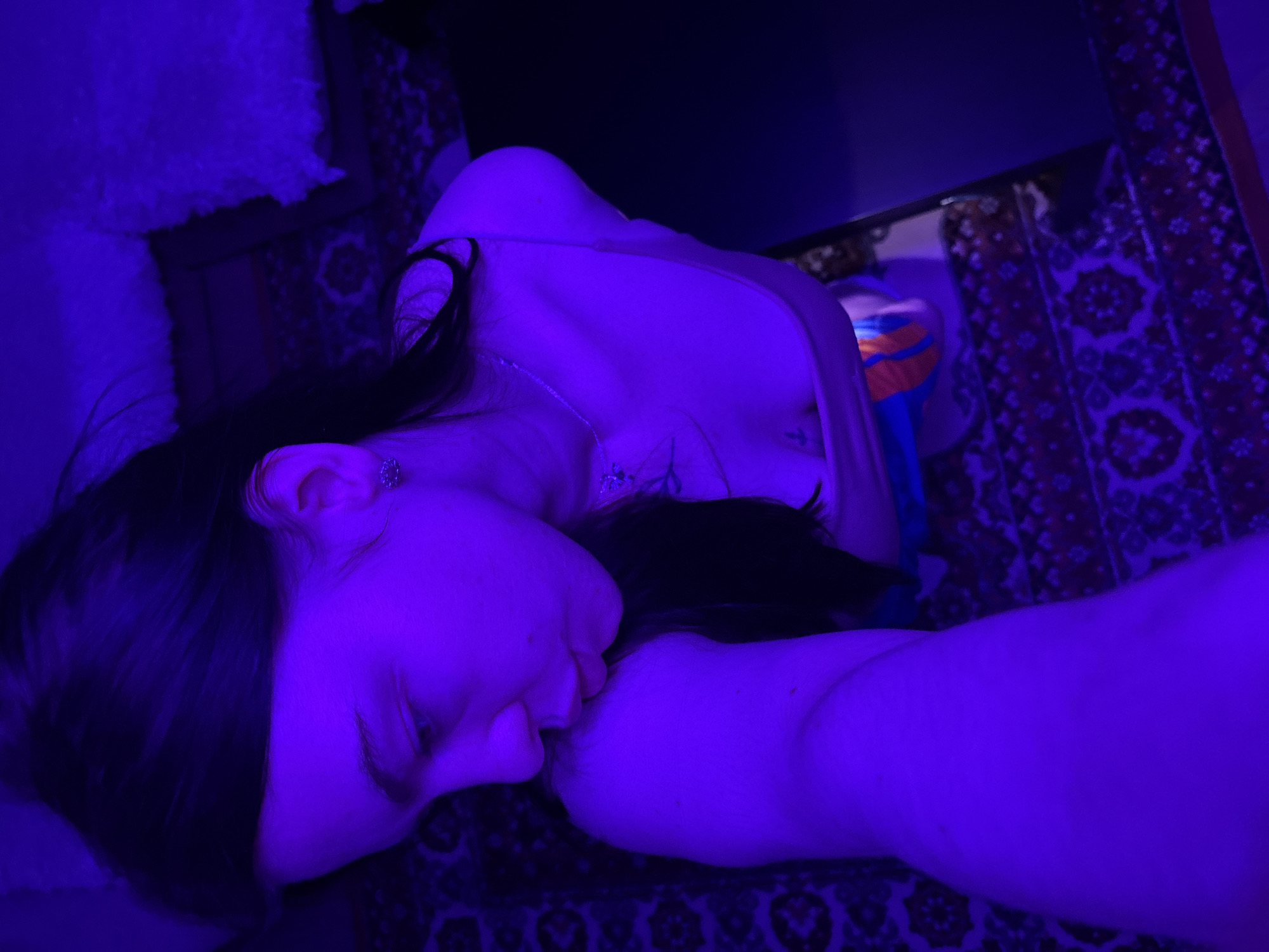 The image depicts a woman lying down on a patterned rug. She has dark hair and is wearing a necklace. The lighting is predominantly blue. A table or shelf is visible in the background, and a fuzzy object is in the upper left corner. The location is purported to be Perm, Russia, though this cannot be independently verified from the image alone.