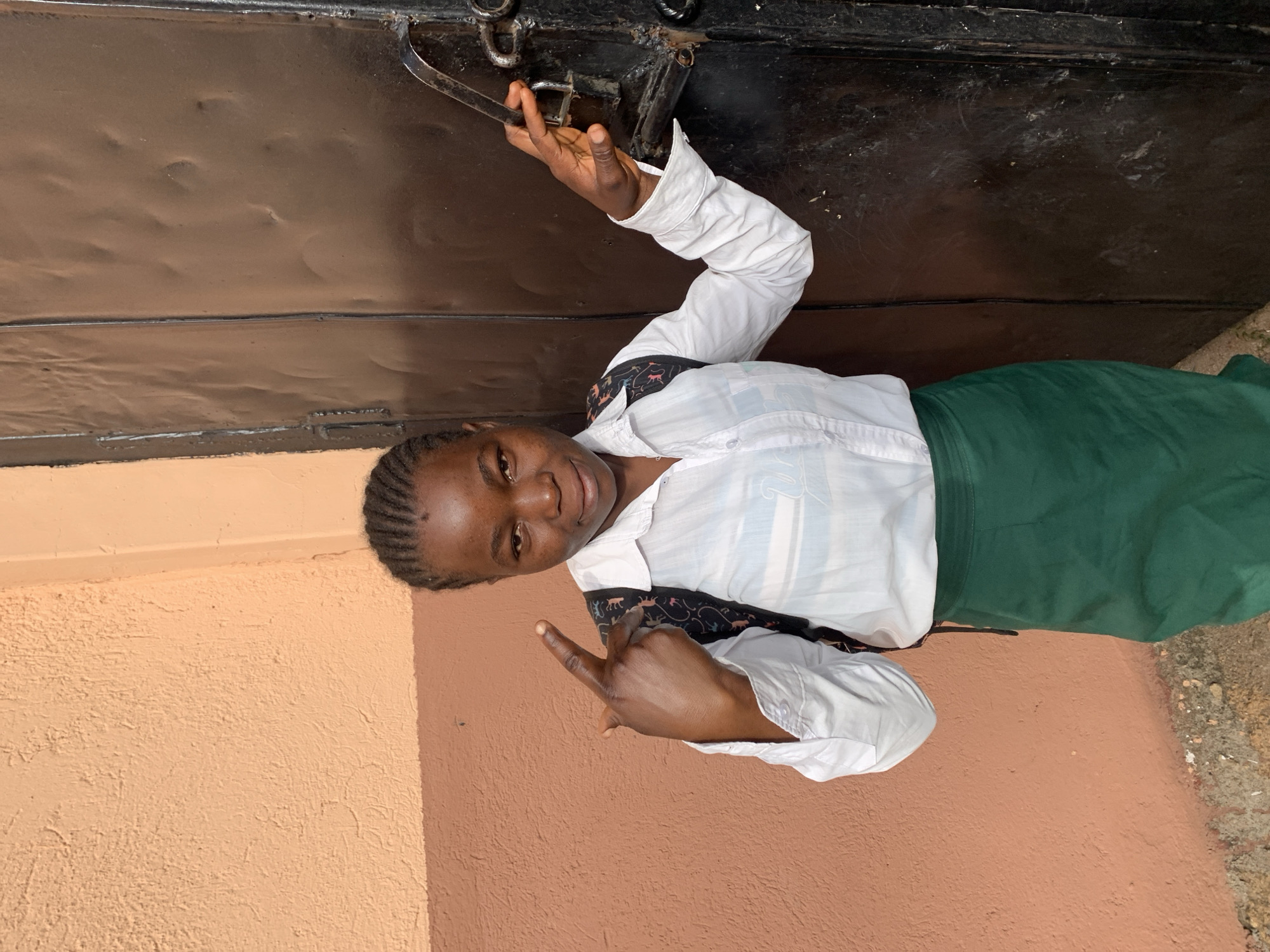 A young woman stands outdoors in Jos, Nigeria, near a building with peach-colored exterior walls and a dark brown door with visible hinges and a latch mechanism. The woman wears a white long-sleeved shirt, a dark vest with a patterned design, and a green skirt. She is making a gesture with her right hand, while her left hand appears to be reaching toward the door's latch. The ground is visible, suggesting an exterior setting.