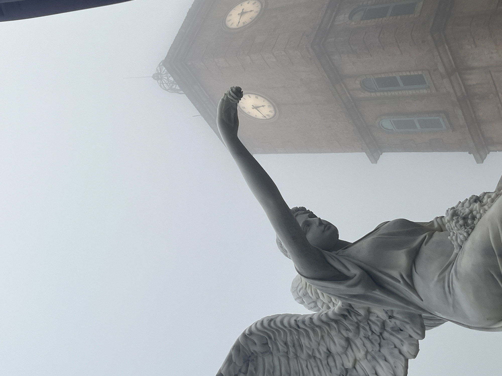 The image depicts a marble statue of an angel with large feathered wings, facing to the right in profile. The angel is seated, holding a bouquet of flowers in its lap and extending its right arm forward, with its hand reaching out and holding what appears to be a single flower. Behind the statue, a tall, stone clock tower with two visible clock faces is shrouded in heavy fog. The tower is made of weathered, brown stone blocks and features arched windows. The clock faces are illuminated, showing Roman numerals. The sky is overcast and filled with thick, uniform fog, obscuring the background and creating a diffused light. The scene is likely located at the Ba Na Hills French Village in Đà Nẵng, Vietnam, a popular tourist destination known for its European-inspired architecture and misty mountain climate. The overall atmosphere is ethereal and somewhat melancholic due to the fog and the solemn pose of the angel statue.