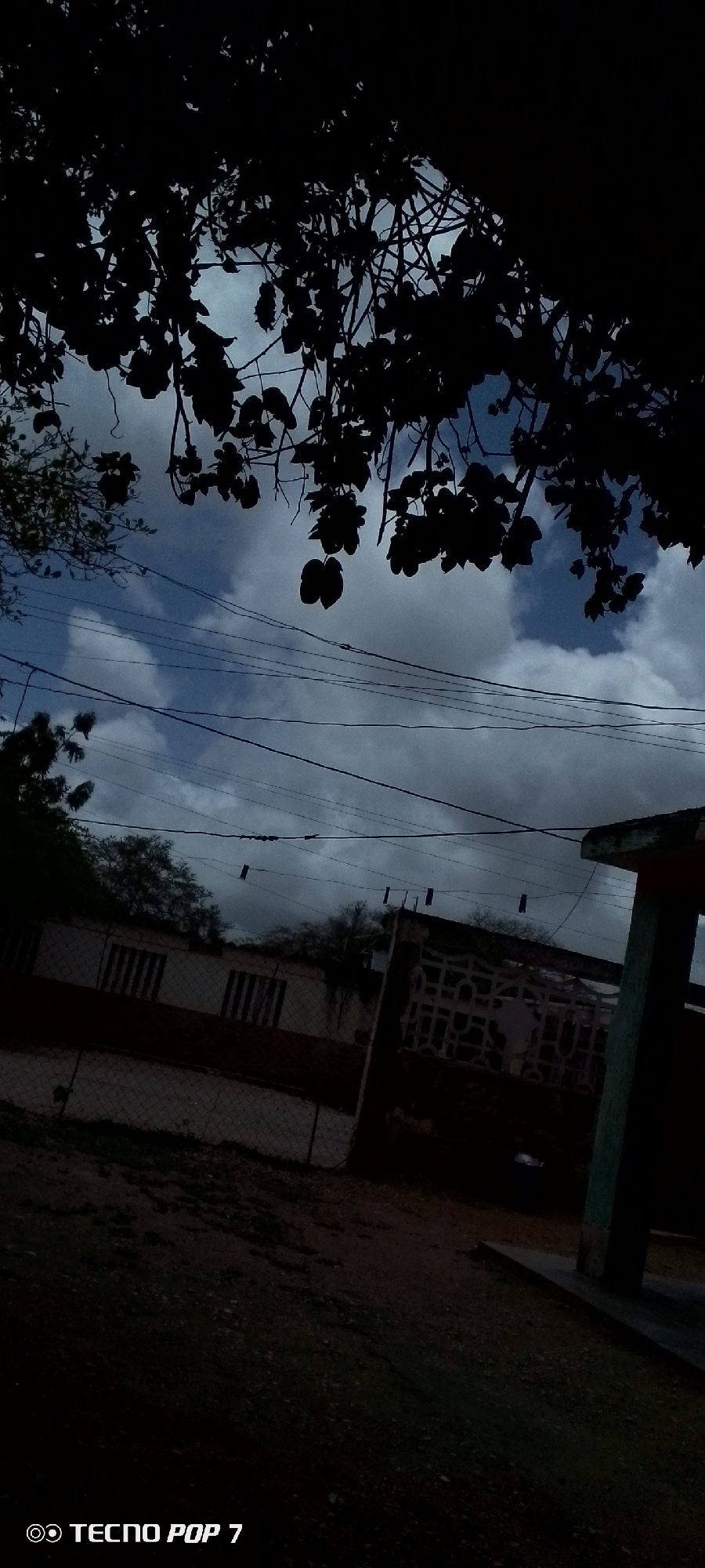 The image captures a scene in Altagracia, Venezuela, under a cloudy sky. In the foreground, dark silhouettes of tree branches and leaves partially obscure the view of the sky, which is a mix of blue and gray clouds. Several electrical wires are strung across the frame, with what appear to be clothespins attached to them.
Below the wires, the middle ground shows a fence and a building. The building has a striped pattern on its facade, suggesting windows or decorative elements. Part of another building, possibly a structure with pillars and a roof, is visible on the right side of the image, and a concrete base extends from it. The ground in the foreground is dark and appears to be covered with small stones and dirt. The overall impression is one of an outdoor, possibly residential or semi-urban, environment. The image has a dark, slightly underexposed quality. The bottom left corner displays the text "TECNO POP 7".