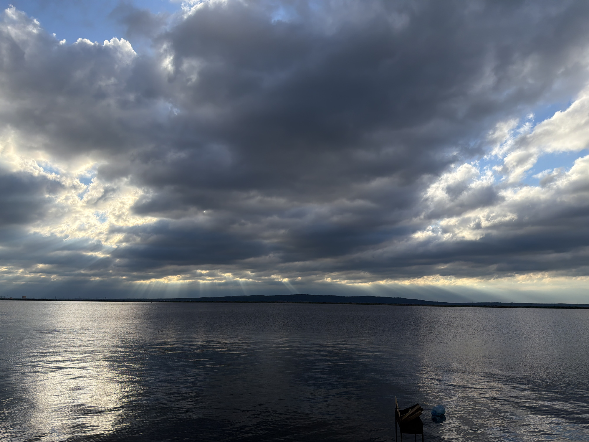 This media file captures a serene seascape under a dramatic, cloudy sky. The scene is dominated by a vast expanse of dark, rippling water reflecting the subdued light. Above, a thick layer of dark, heavy clouds stretches across the horizon, interspersed with patches of bright, illuminated sky. Beams of sunlight pierce through the clouds, creating a striking crepuscular effect, casting rays of light downwards onto the water.
The horizon is marked by a distant, low-lying landmass, possibly a shoreline, silhouetted against the bright sky. The time of day appears to be late afternoon or early evening, given the quality of light and the dramatic cloud formations often associated with the approach of sunset or a storm. The weather is cloudy, with moments of sunshine breaking through, creating a dynamic and somewhat moody atmosphere.
In the foreground, to the right, a small grill or barbecue setup is partially visible at the edge of the water, along with a blue floating object. These human-made items suggest a recreational setting or a place where people might gather. There are no visible people or text in the image. The overall emotion conveyed is one of calm and vastness, with a touch of awe inspired by the natural spectacle of the sky and water. The location is identified as Chaikovsky, Russia.