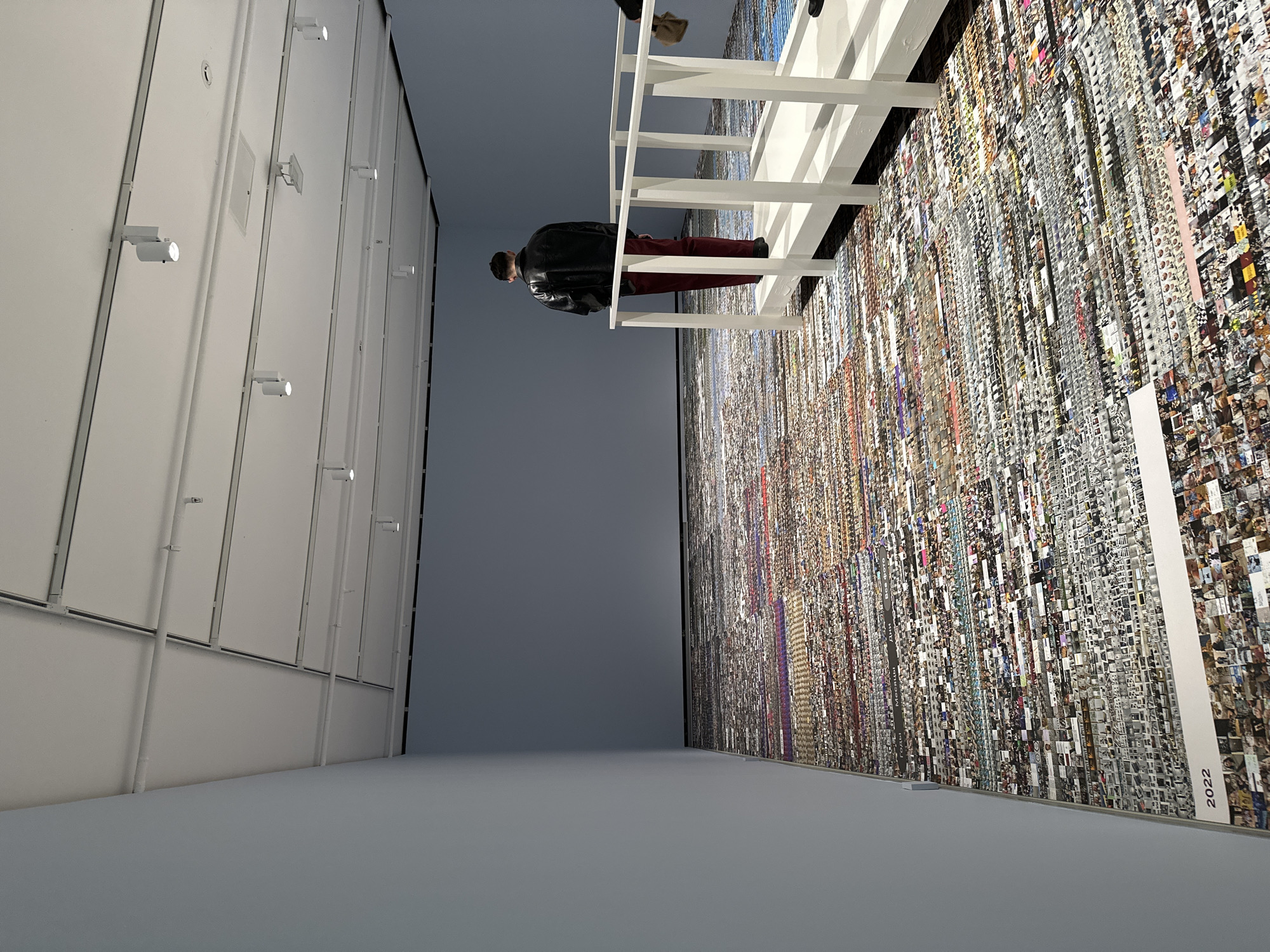 The image depicts an indoor art gallery space. A tall wall is covered with numerous small photos arranged in rows and columns, creating a mosaic effect. A person, presumably an art viewer, stands on a white, elevated platform with railings, observing the artwork. The person is wearing a black jacket and red trousers. The left side of the image features a white wall with vertical panels, each illuminated by a spotlight. The date "2022" is visible at the bottom right corner of the wall covered with photos. The overall impression is of an art exhibition, possibly a large-scale installation using photographic elements. Moscow, Russia cannot be confirmed from this picture.