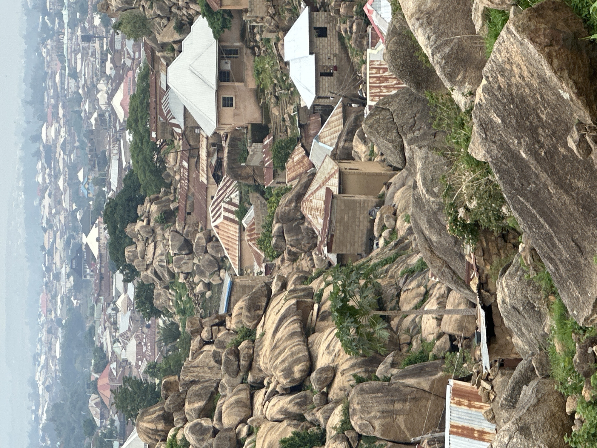 This image presents an elevated view of an urban settlement integrated with a rugged, rocky landscape, characteristic of Jos, Plateau State, Nigeria. The foreground and midground are dominated by numerous large, weathered granite boulders and outcrops, forming natural terraces and crevices.
Scattered amongst these rock formations are various residential structures. These dwellings are primarily constructed from stone blocks or rendered walls, and feature a mix of corrugated metal roofs. Some roofs display significant rust and deterioration, indicating age, while others appear newer with brighter, less corroded silver sheeting. Architectural styles vary, but most are single or two-story buildings. Visible details on some structures include small rectangular windows and doorways. Paths or rudimentary staircases, often carved into or built upon the rock, connect some of the houses.
Sparse vegetation, including small trees, shrubs, and patches of grass, grows in the crevices and soil pockets between the boulders and around the structures. A distinct palm-like tree stands prominently in the lower-midground among the rocks.
In the background, a dense urban area extends into a hazy distance, suggesting a larger city. This background cityscape features a multitude of residential and possibly commercial buildings with diverse roof types, appearing somewhat indistinct due to atmospheric haze or distance.
No individuals or specific human actions are discernible within the frame. The overall lighting suggests an overcast or hazy sky, contributing to a soft, diffuse illumination across the scene.